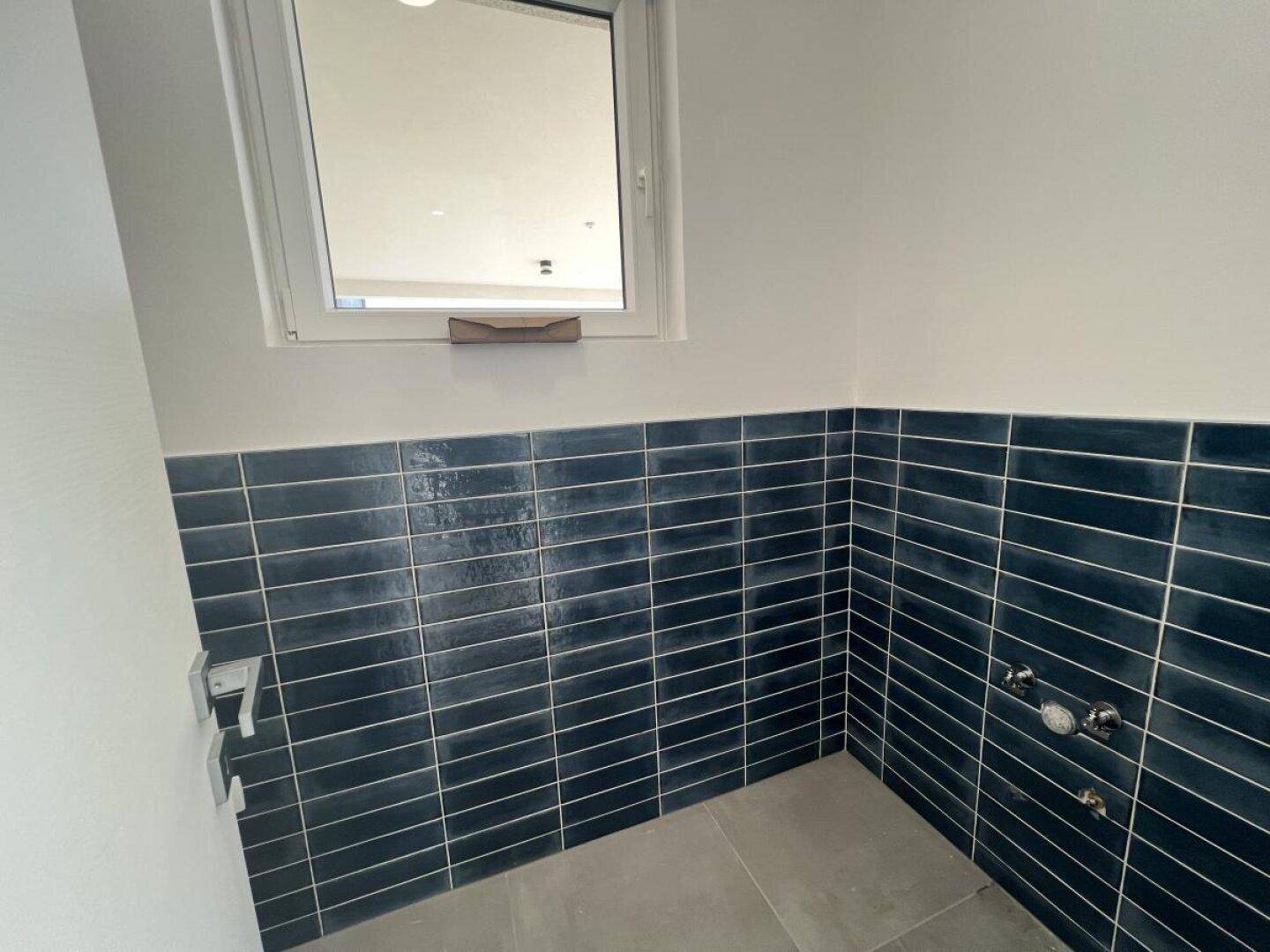 Kleiner Raum mit blauen Wandfliesen, Fenster und Anschlüssen für Sanitäranlagen.