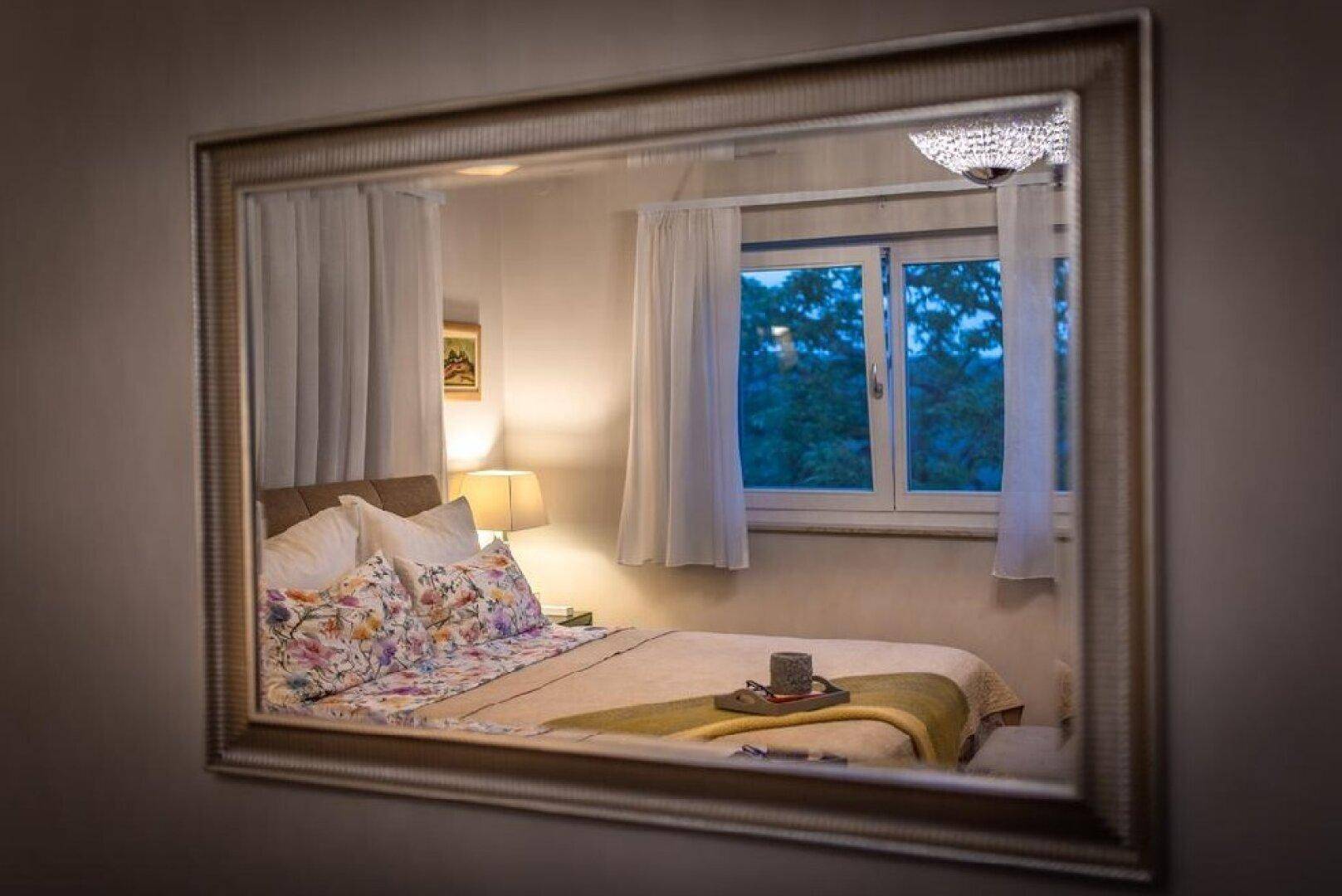 Schlafzimmer mit floraler Bettwäsche, Nachttischlampe und Fenster, im Spiegel reflektiert.