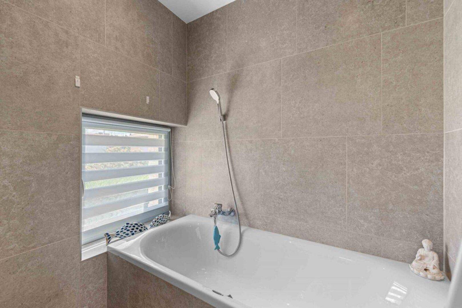 Helles Badezimmer mit Badewanne und Fenster, ausgestattet mit modernen Fliesen.