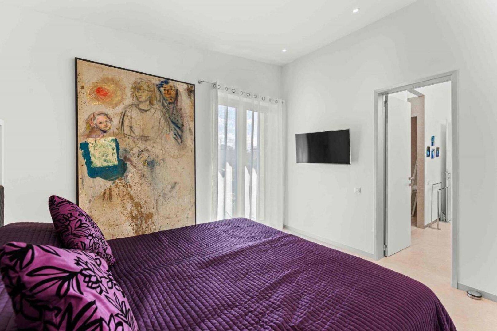 Helles Schlafzimmer mit Doppelbett, Wandfernseher und Zugang zu einem Außenbereich.