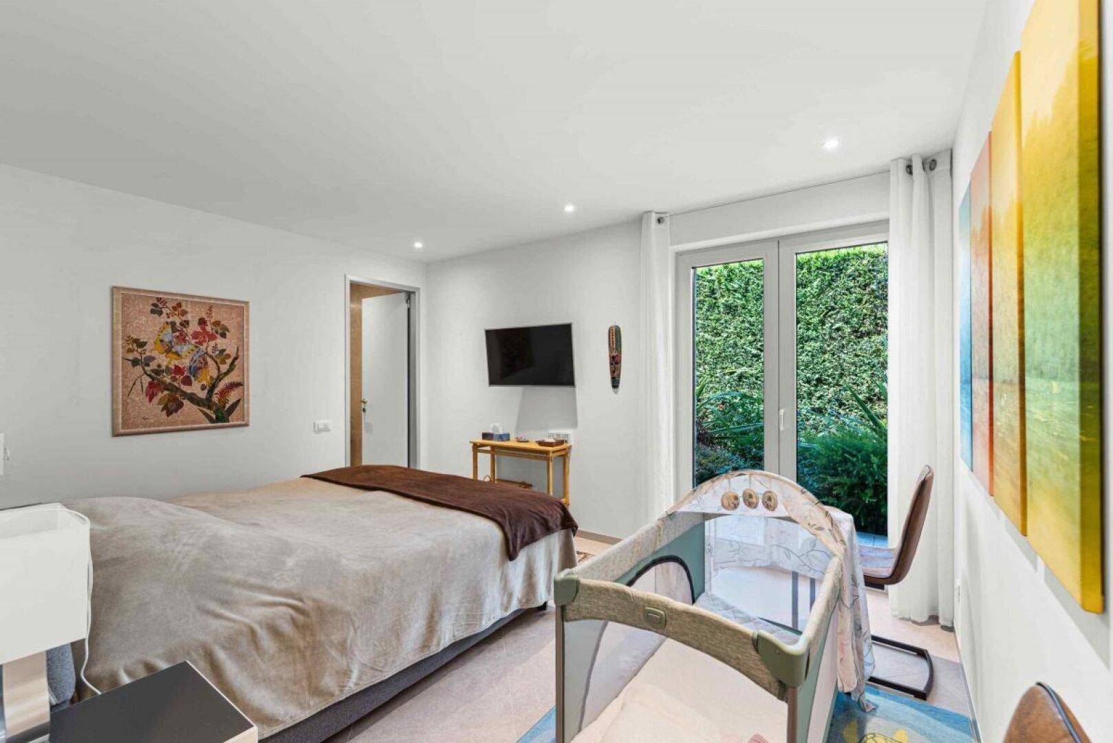 Helles Schlafzimmer mit Doppelbett und Babybett, mit Blick auf eine grüne Hecke.