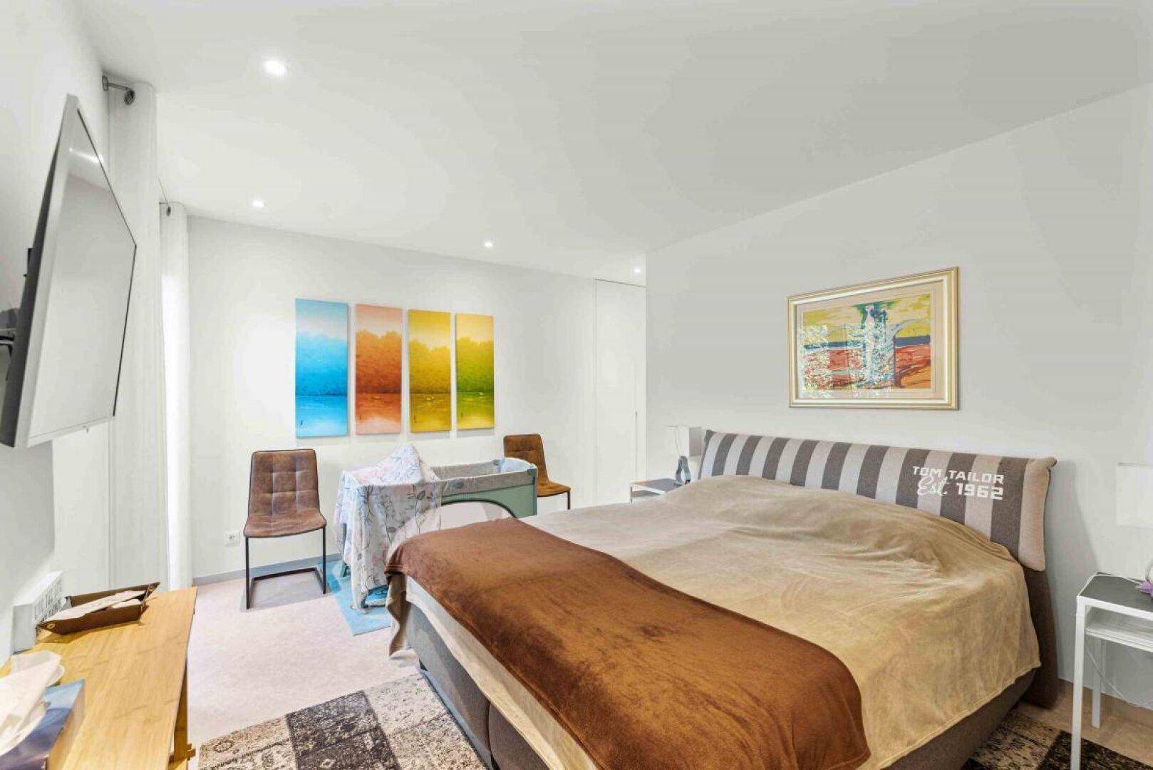 Geräumiges Schlafzimmer mit Doppelbett, Babybett und Wandfernseher, hell und modern eingerichtet.