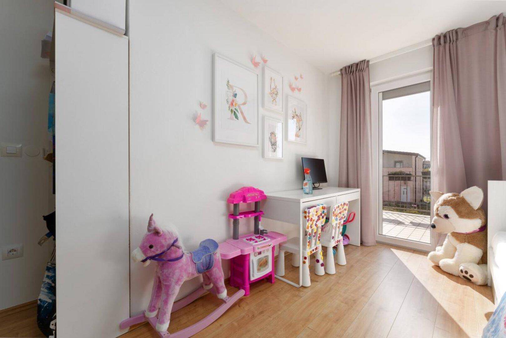 Helles Kinderzimmer mit Schreibtisch, Spielzeug und großem Fenster zum Balkon.