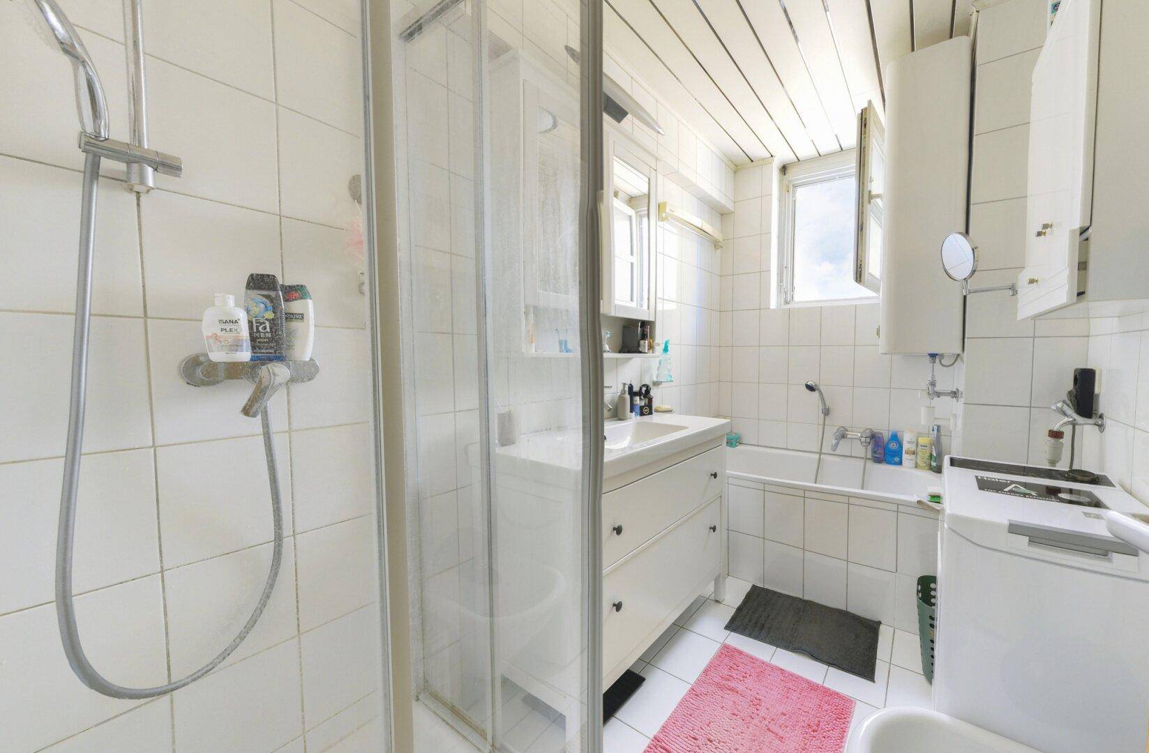 Helles Badezimmer mit Dusche, Badewanne, Waschbecken und Fenster, komplett gefliest.