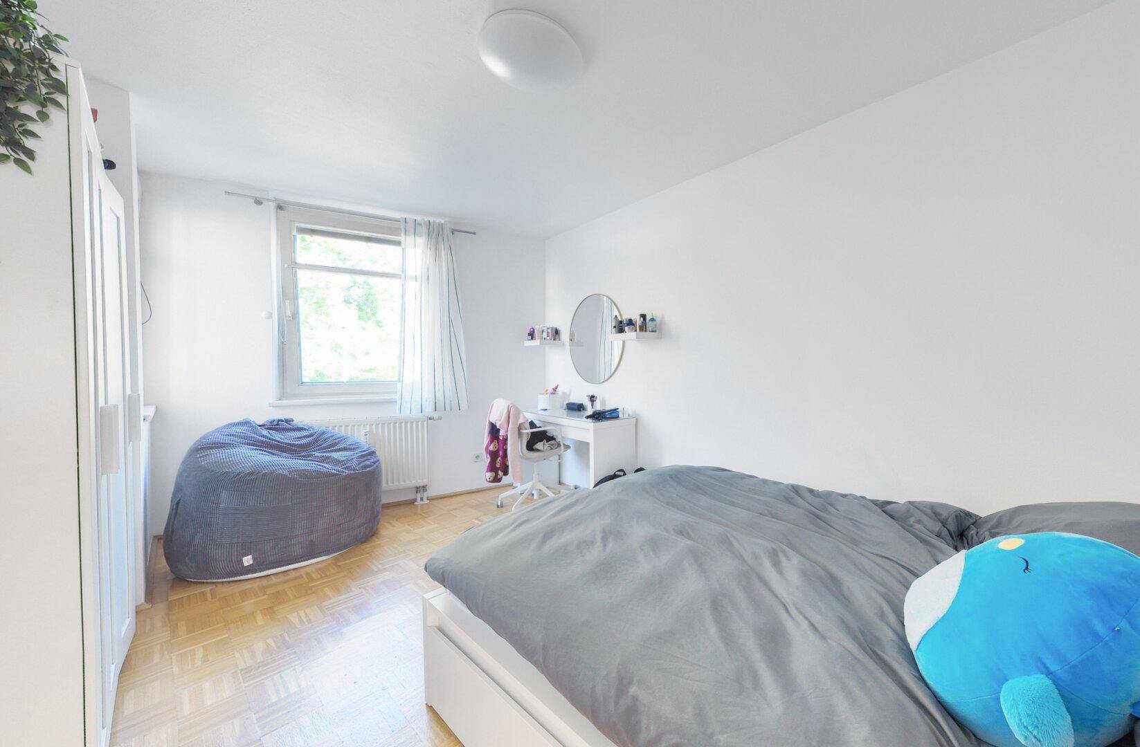 Freundliches Schlafzimmer mit Bett, Schreibtisch und Fenster, das viel Tageslicht hereinlässt.