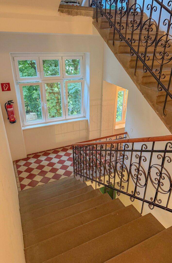 Historisches Treppenhaus mit schmiedeeisernem Geländer, großen Fenstern und Blick ins Grüne.