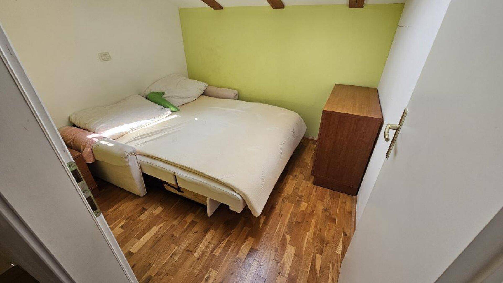 Kleines Schlafzimmer mit Holzboden, Bett und Kommode, akzentuiert durch eine grüne Wand.