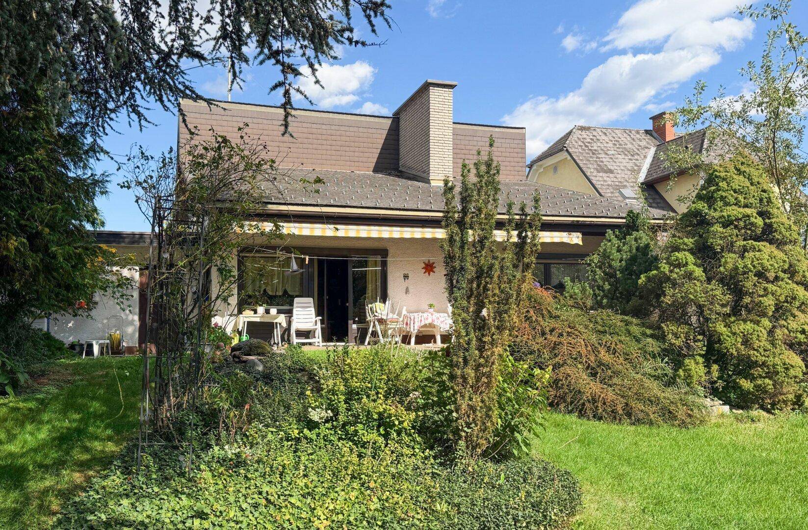 Rückseite des Hauses mit gepflegtem Garten, großer Terrasse und Markise unter blauem Himmel.