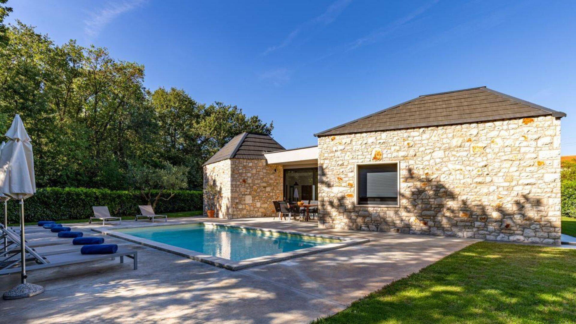 Weitläufiger Poolbereich mit Sonnenliegen und der Steinfassade der modernen Villa im Hintergrund.