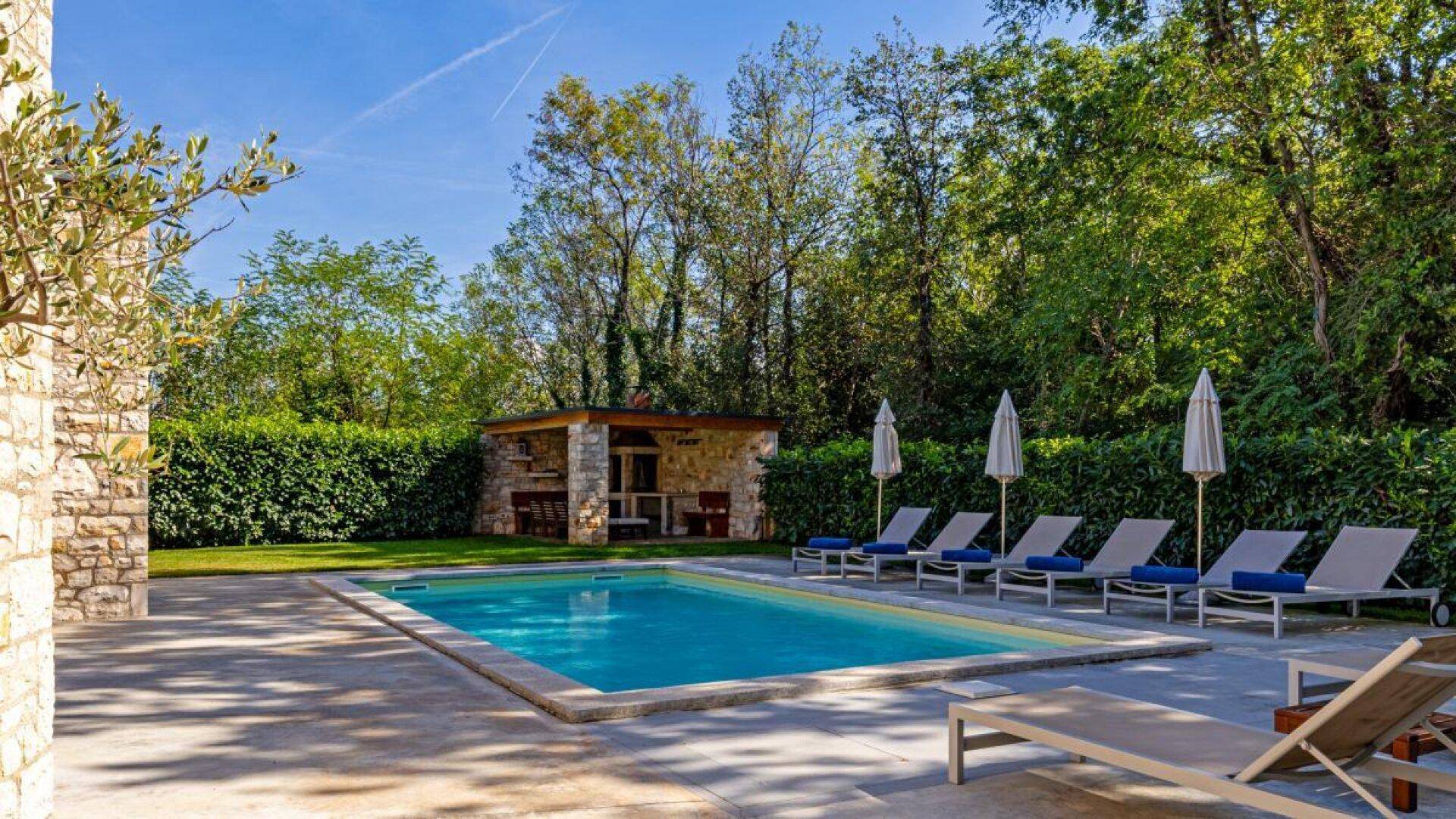 Entspannender Poolbereich mit Sonnenliegen und einer überdachten Außenküche, umgeben von grüner Hecke.