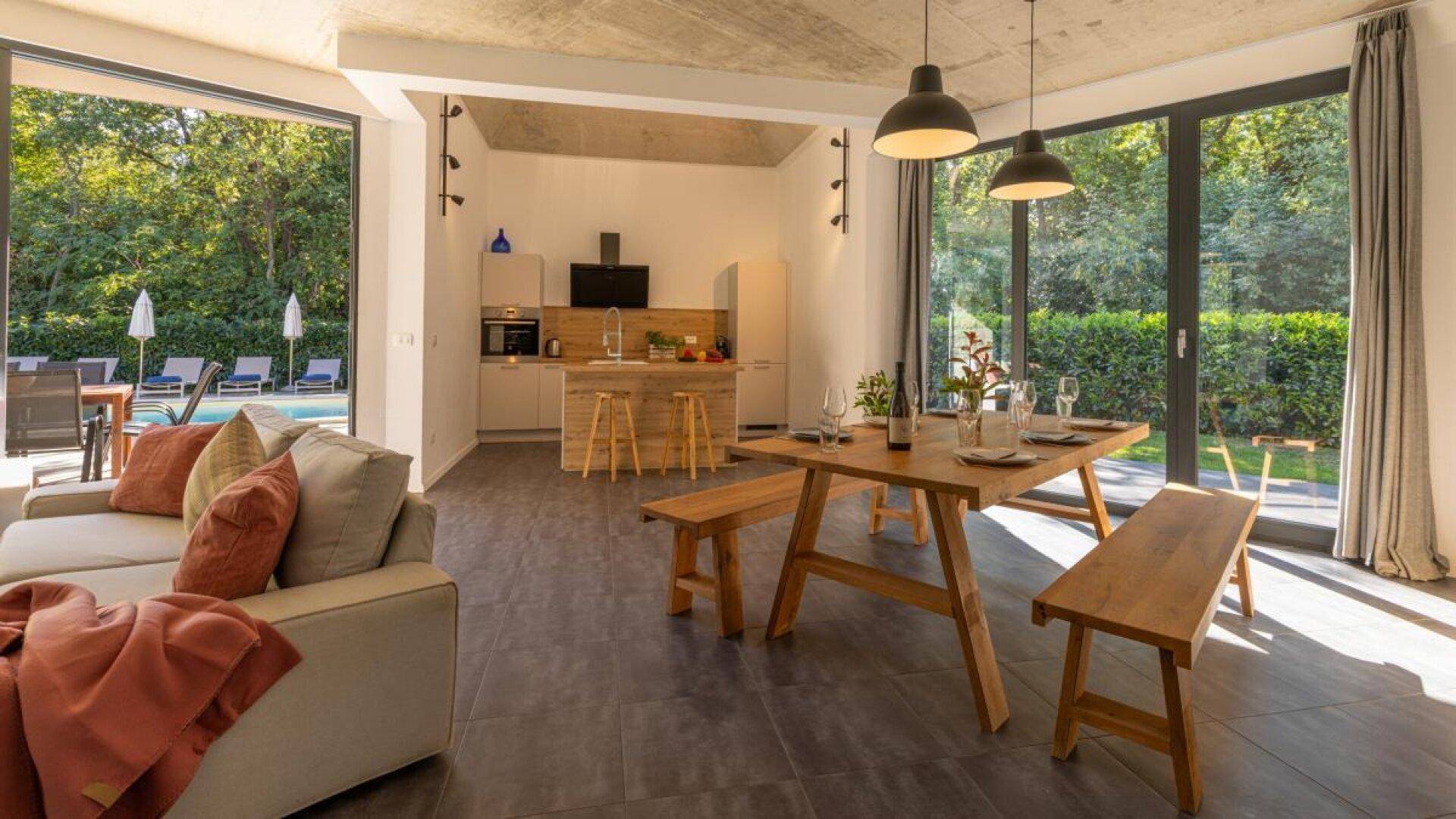Geräumiger Essbereich mit Holztisch und Bänken, offener Küche und Zugang zum Poolbereich.
