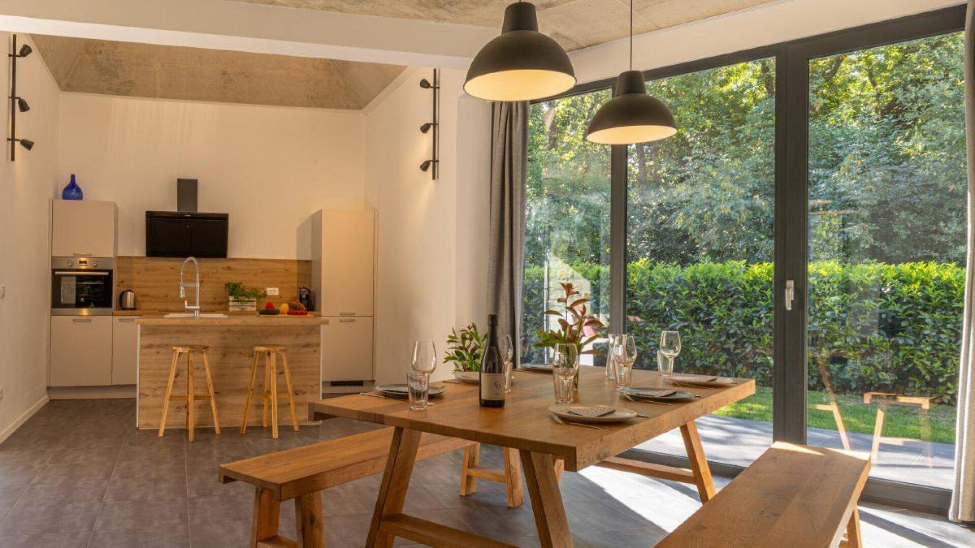 Moderner Essbereich mit Holztisch und Bänken, offener Küche und großen Fenstern zum Garten.