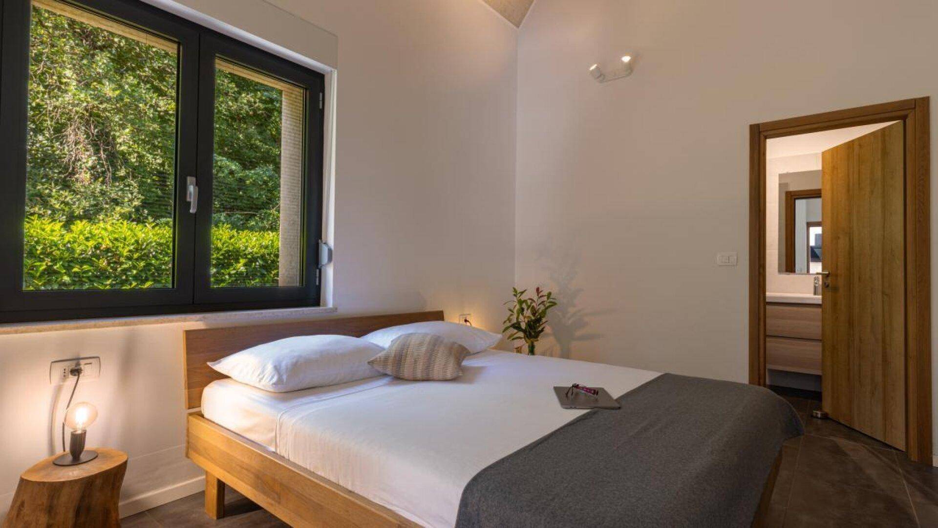 Helles Schlafzimmer mit Holzbett, Nachttisch und Fenster mit Blick ins Grüne.
