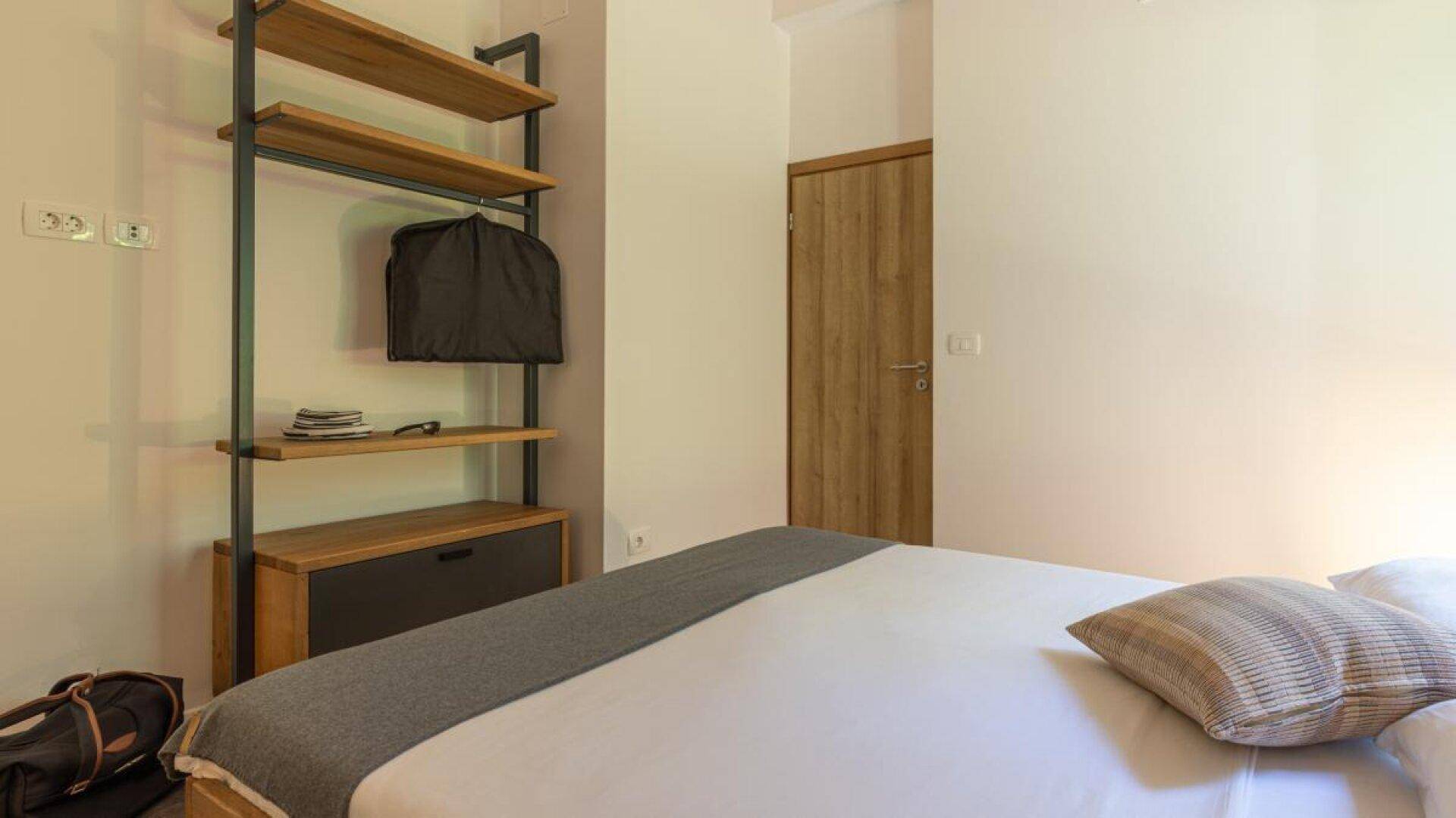Schlafzimmer mit Holzbett und offenem Regal für Kleidung und persönliche Gegenstände.