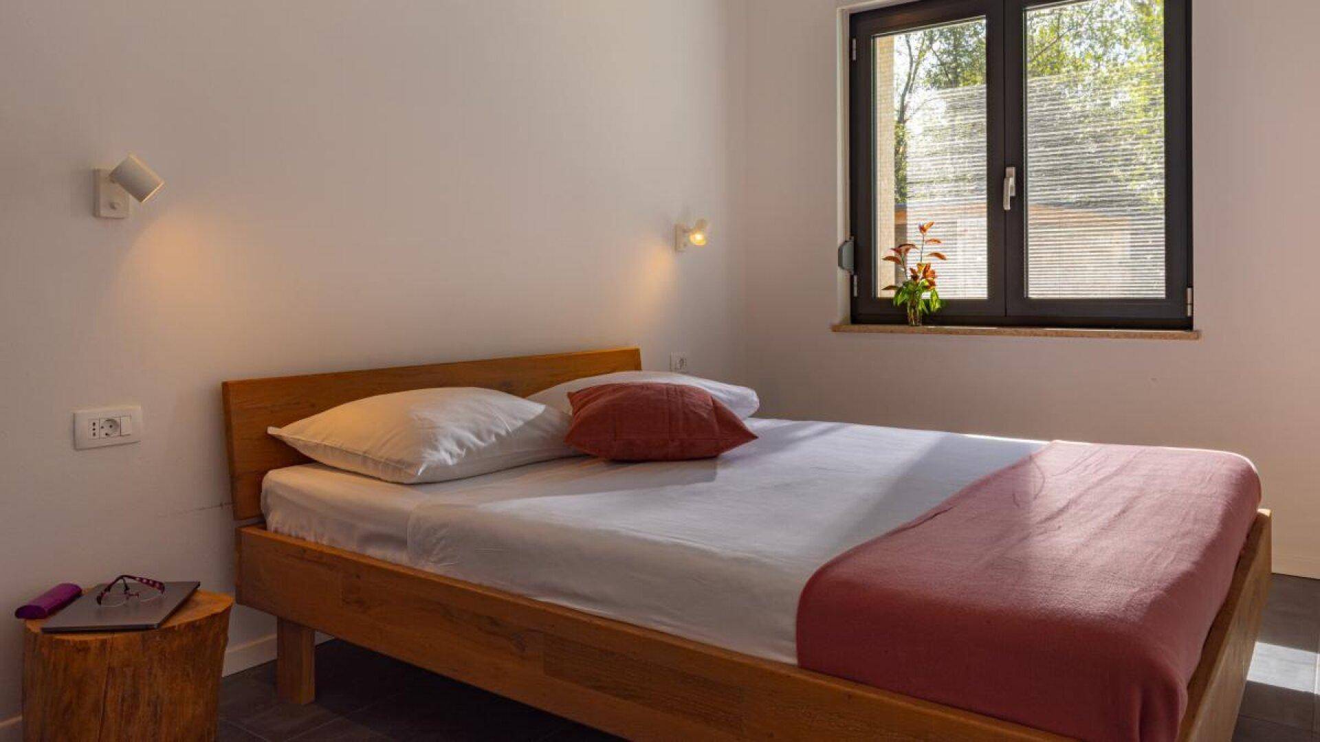 Gemütliches Schlafzimmer mit Holzbett, Nachttisch und Fenster mit Jalousien und Blick ins Grüne.