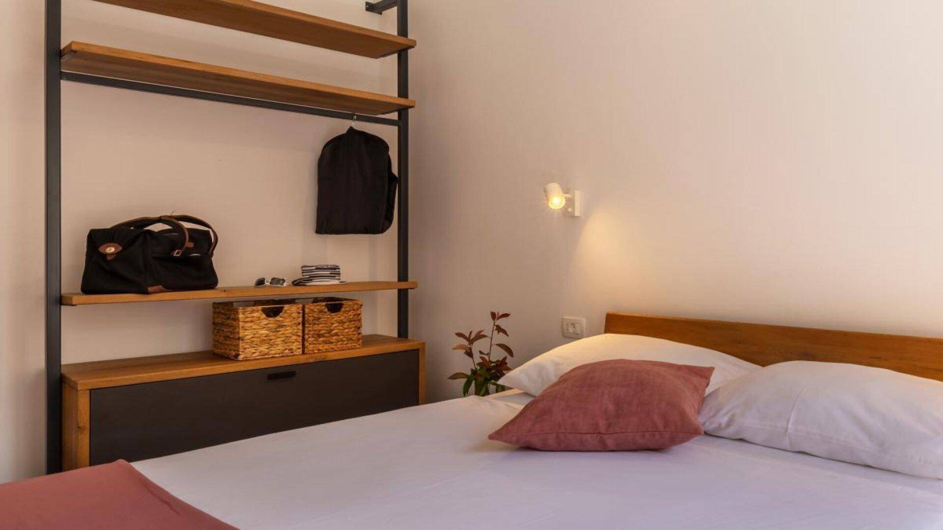 Detailansicht des Schlafzimmers mit Holzbett, Kissen und offenem Regal für persönliche Gegenstände.