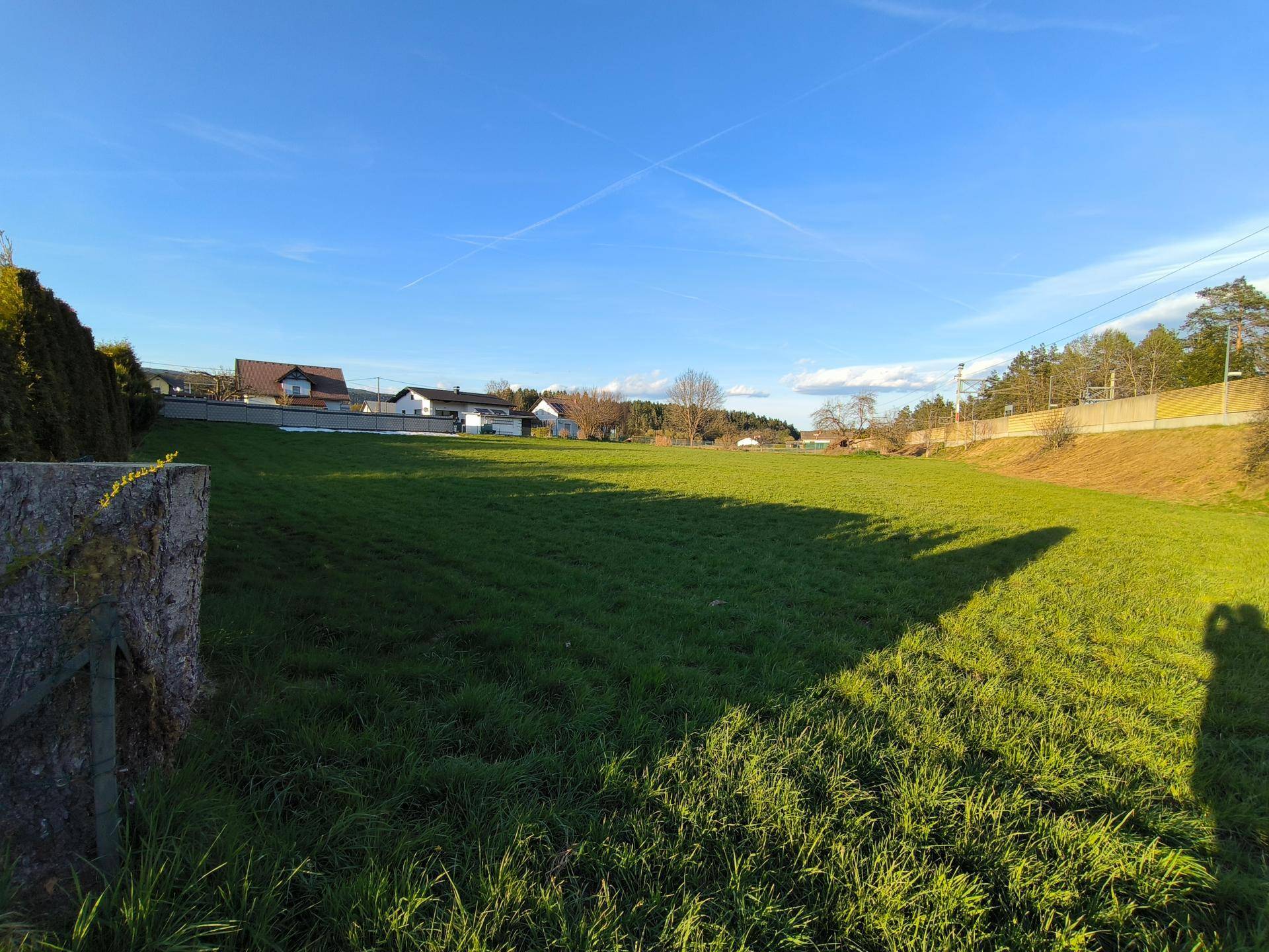 Sonniges, offenes Grundstück mit grüner Wiese, Häusern in der Ferne und einem klaren blauen Himmel.
