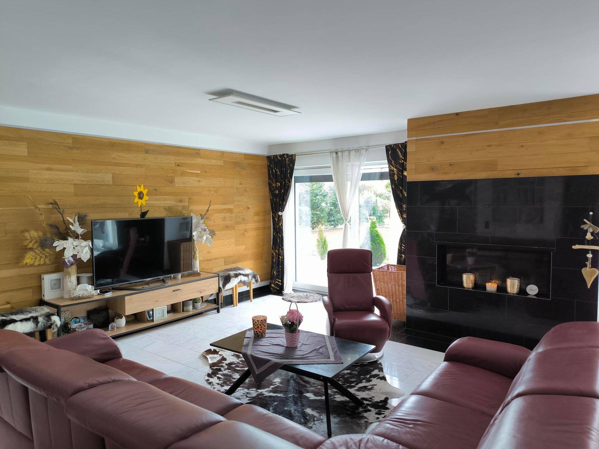 Gemütliches Wohnzimmer mit Ledersofas, Holzwand und modernem Kamin, hell durch große Fenster.