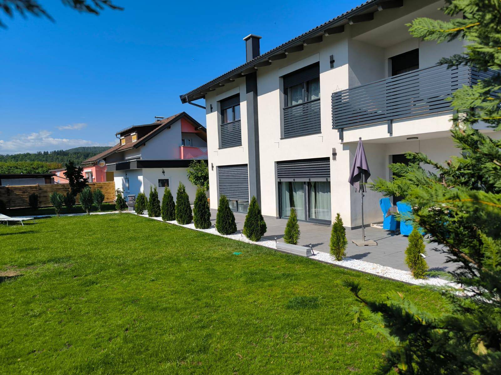 Weitläufiger Gartenbereich mit Rasenfläche, kleiner Terrasse und moderner Hausfassade unter blauem Himmel.