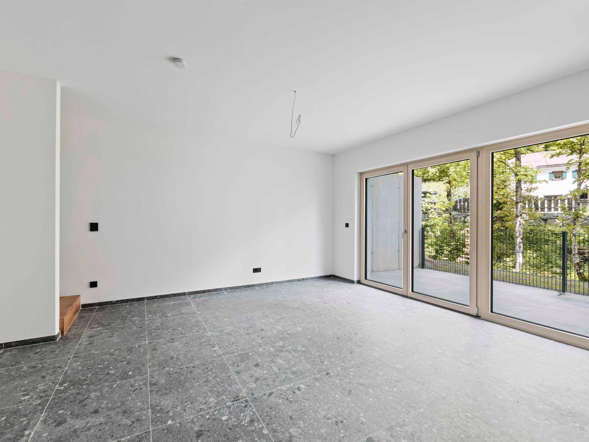 Heller Innenraum mit dunklem Fliesenboden und großen Fenstern mit Zugang zur Terrasse.