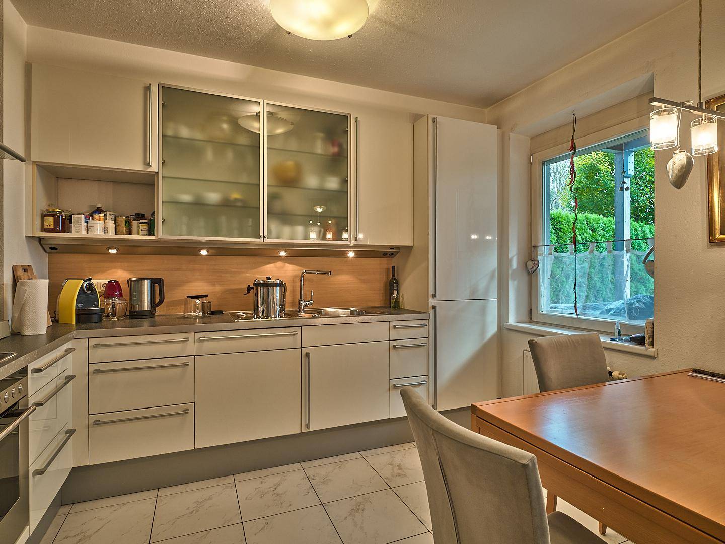 Moderne Küche mit weißen Schränken, Fliesenboden und Essbereich am Fenster.
