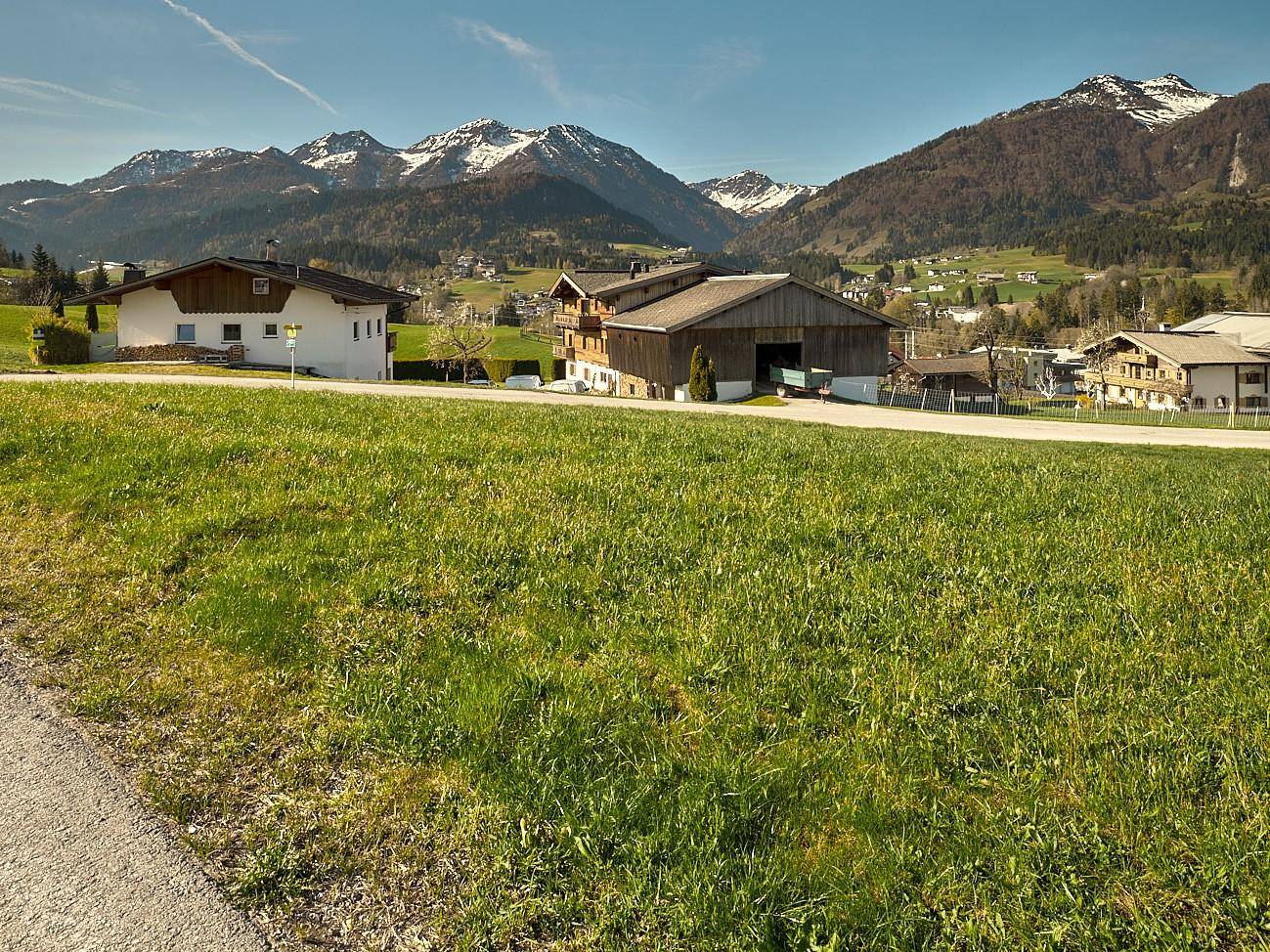 Grüne Wiese mit Häusern und schneebedeckten Bergen im Hintergrund unter blauem Himmel und Sonnenschein.