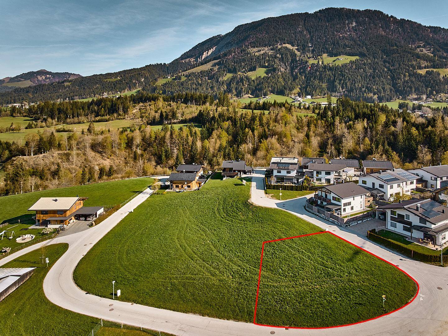 Luftaufnahme eines Baugrundstücks mit roter Markierung inmitten einer modernen Siedlung und Bergkulisse.