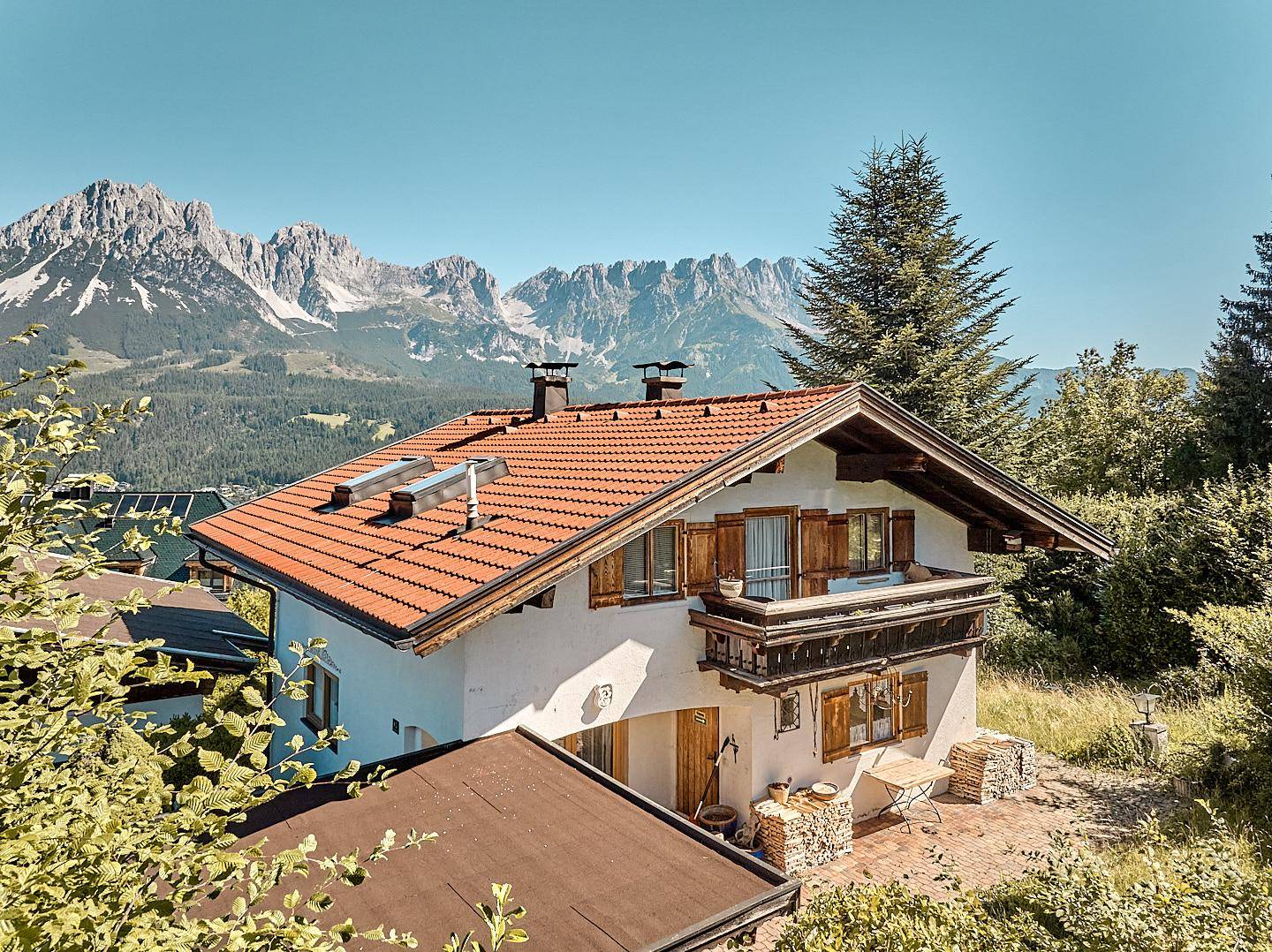 Weiß verputztes Haus mit rotem Ziegeldach und Balkon, umgeben von grüner Landschaft und Bergen.