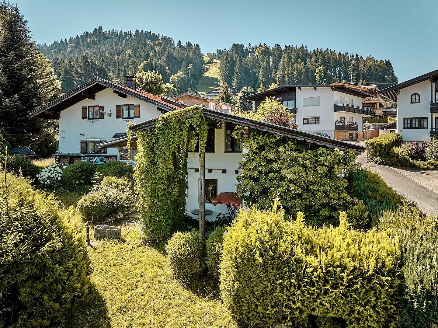 Charmantes Haus mit üppiger grüner Bepflanzung und weiteren Gebäuden im Hintergrund einer Berglandschaft.