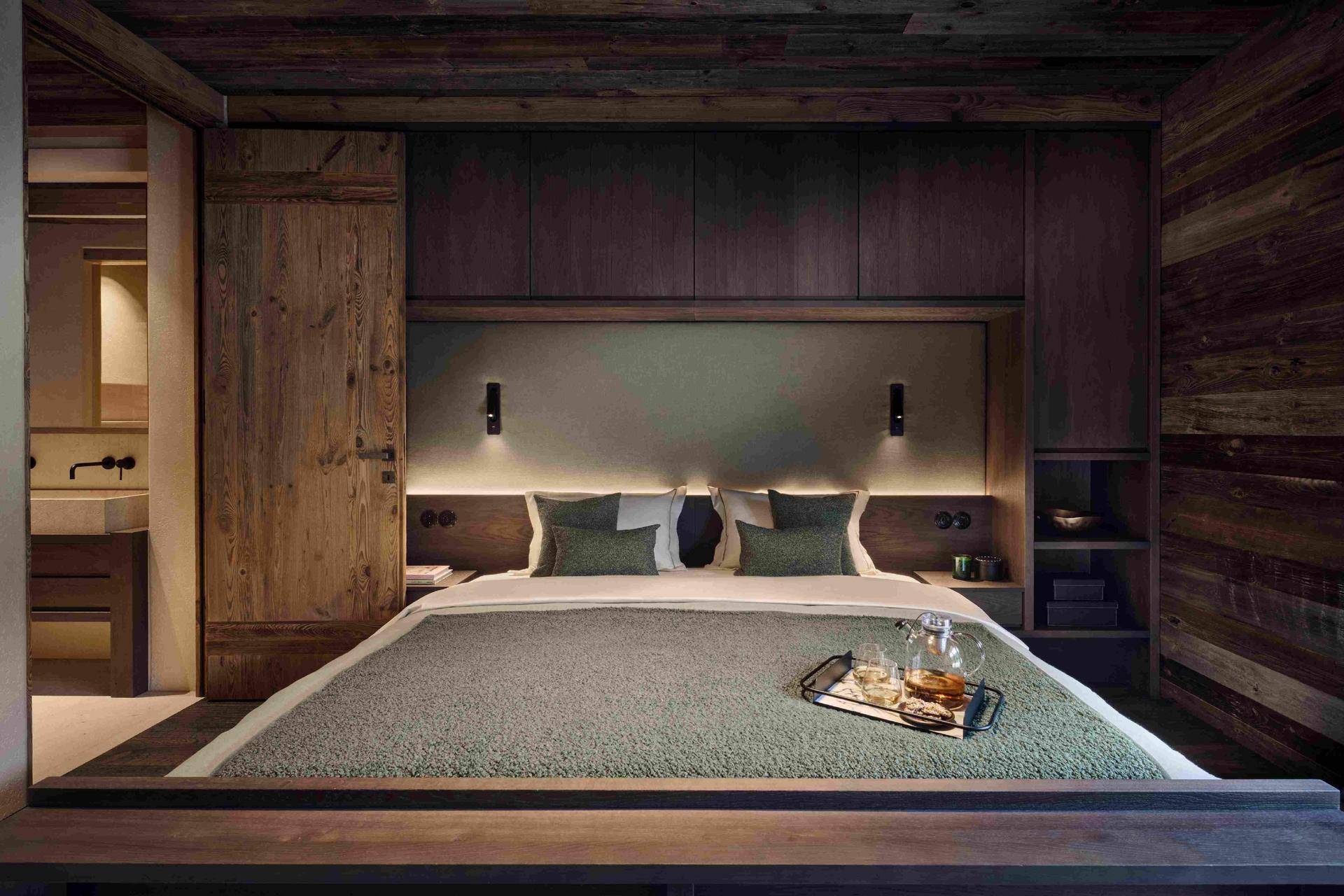 Gemütliches Schlafzimmer mit Holzwänden, integriertem Kopfteil, indirekter Beleuchtung und Zugang zum Bad.
