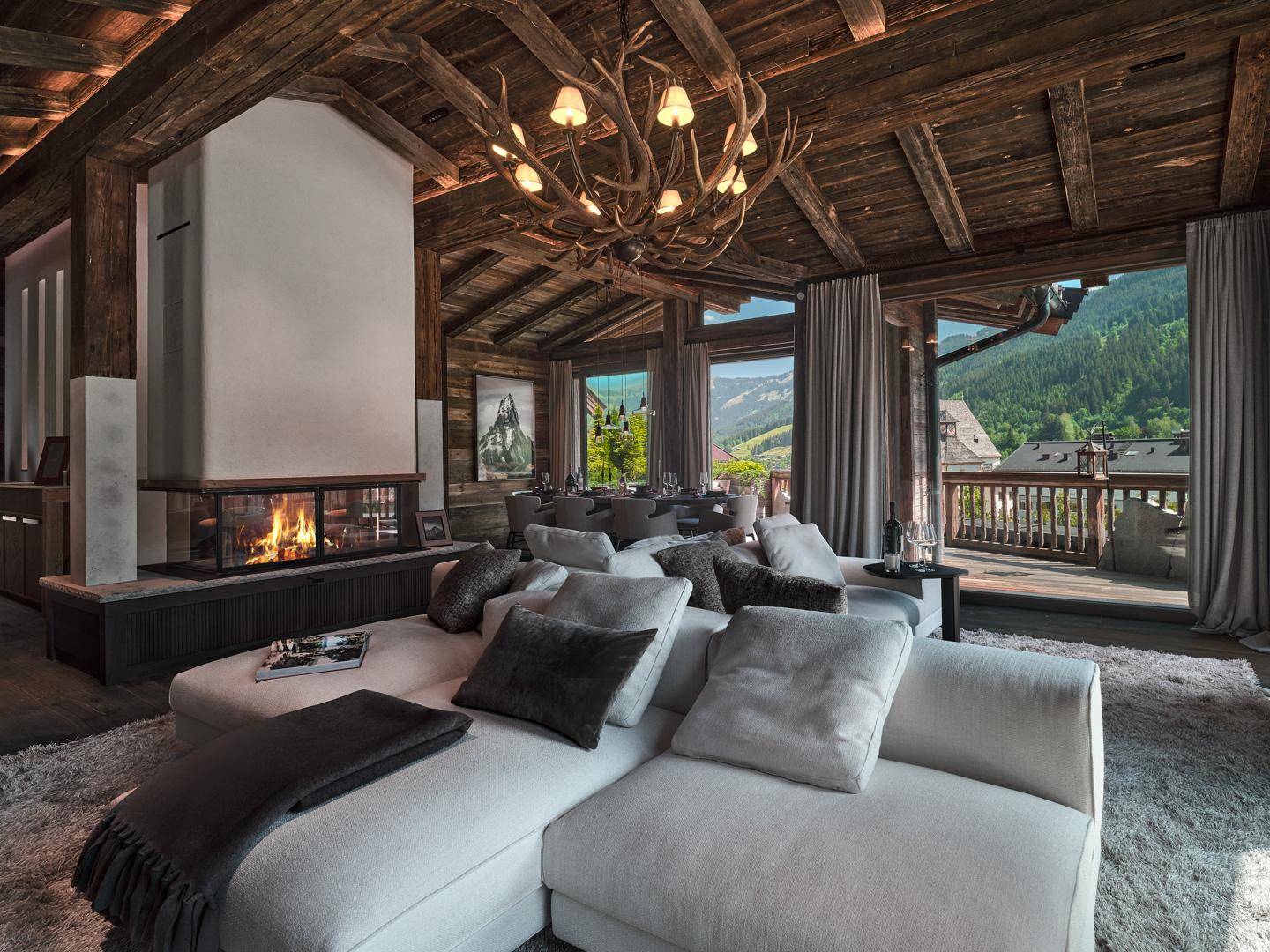 Gemütliches Wohnzimmer im alpinen Stil mit Kamin, Holzbalken, Geweihleuchter und Bergblick.