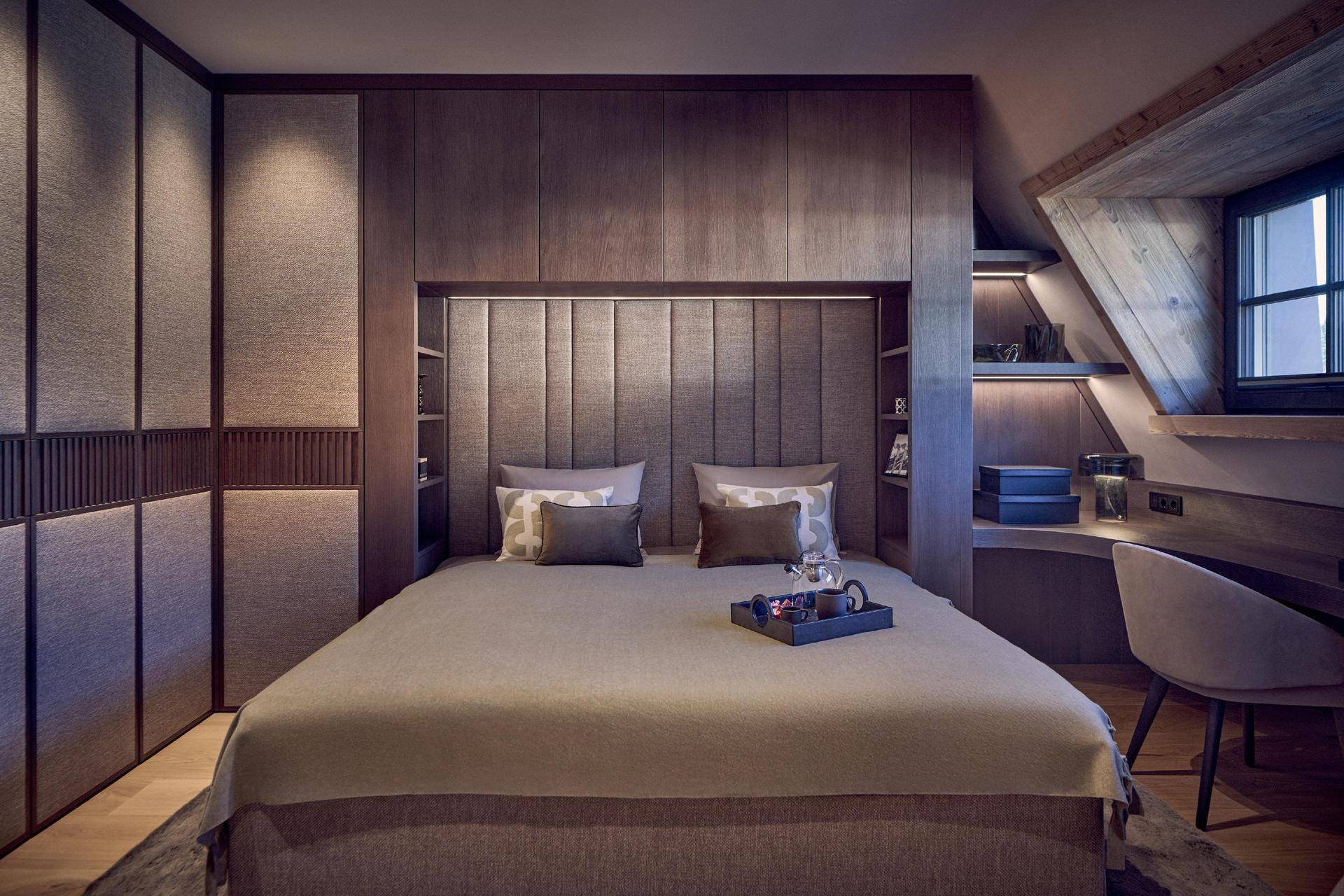 Schlafzimmer mit Holzeinbauten, gemütlichem Bett und Schreibtisch unter der Dachschräge mit Fenster.
