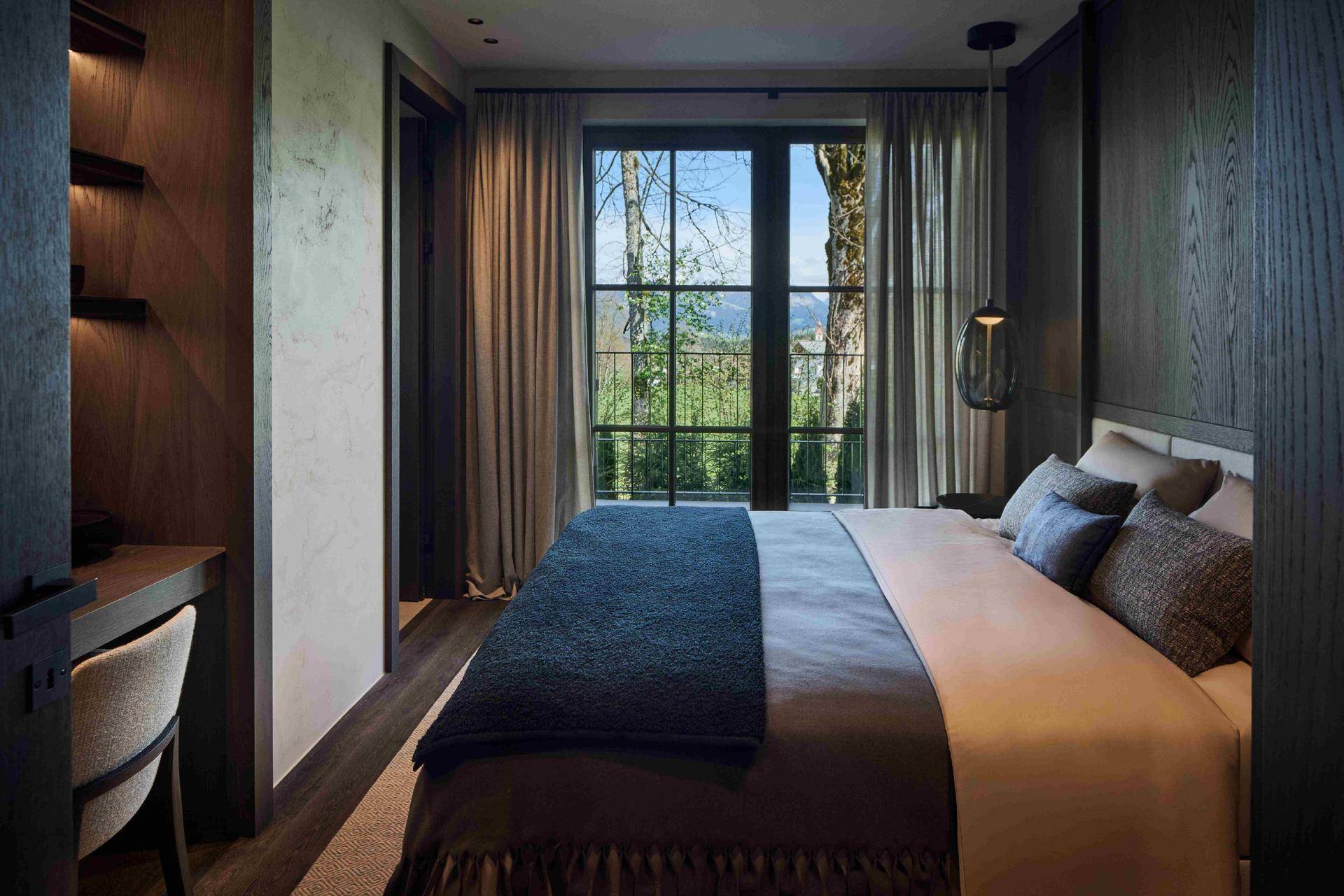 Gemütliches Schlafzimmer mit Holzwänden, großem Bett und Blick ins Grüne durch die Fenster.