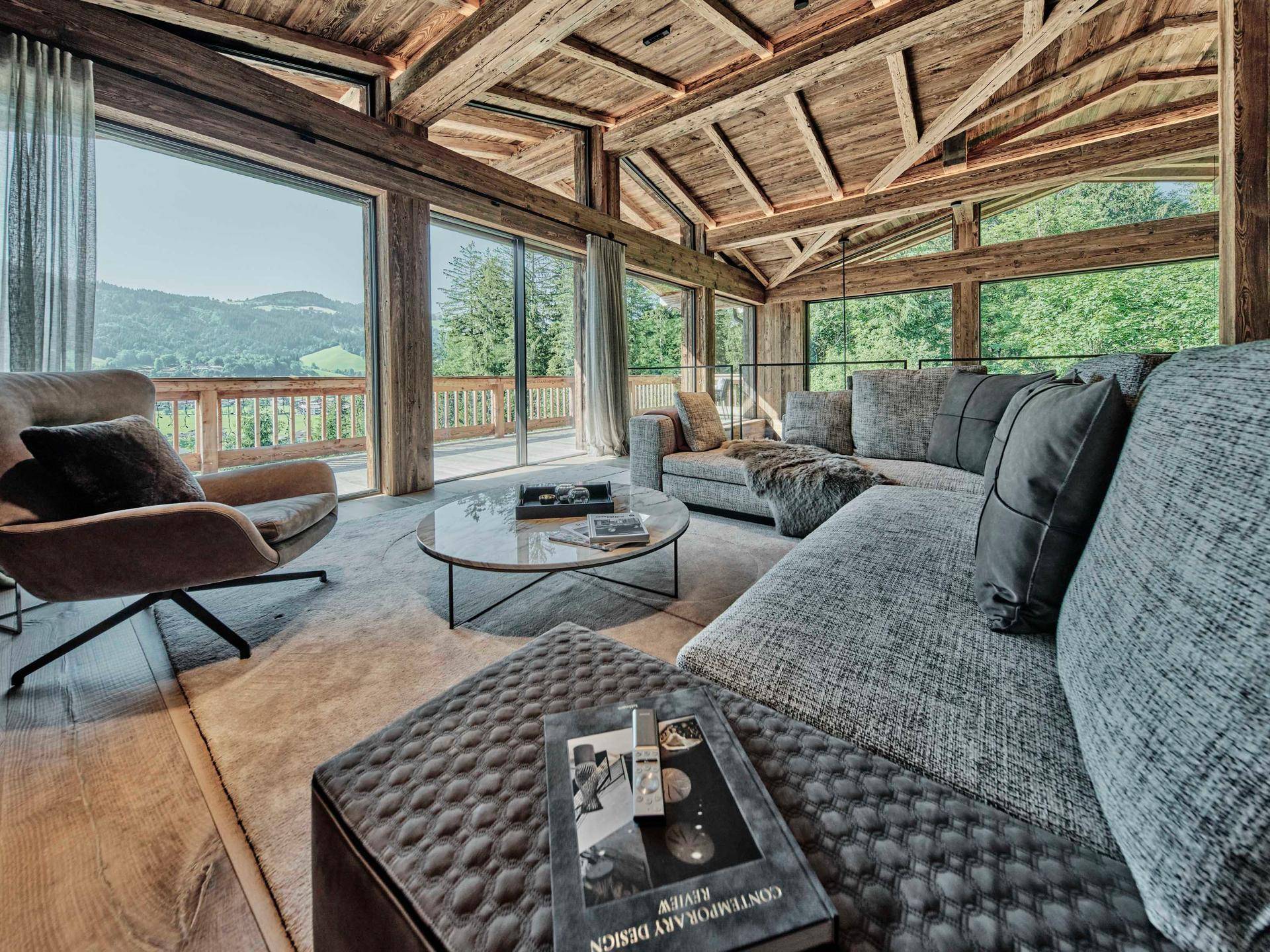 Helles Wohnzimmer mit großzügiger Sofalandschaft, Holzbalken und raumhohen Fenstern mit Blick ins Grüne.