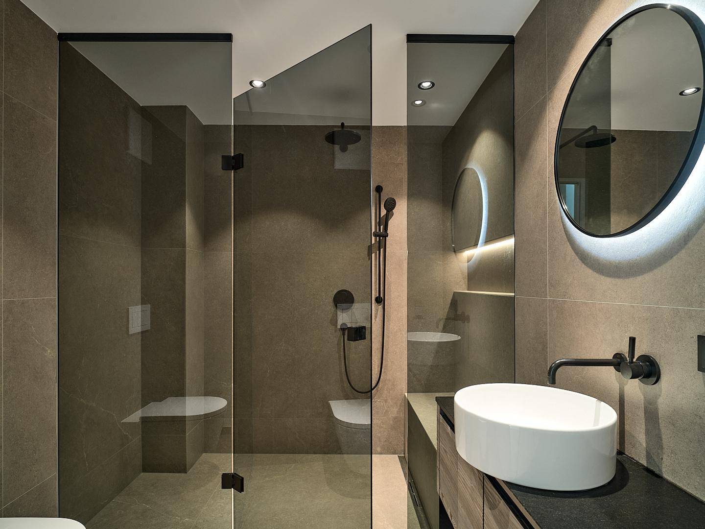 Modernes Badezimmer mit dunklen Fliesen, begehbarer Dusche und beleuchtetem Rundspiegel über dem Waschbecken.