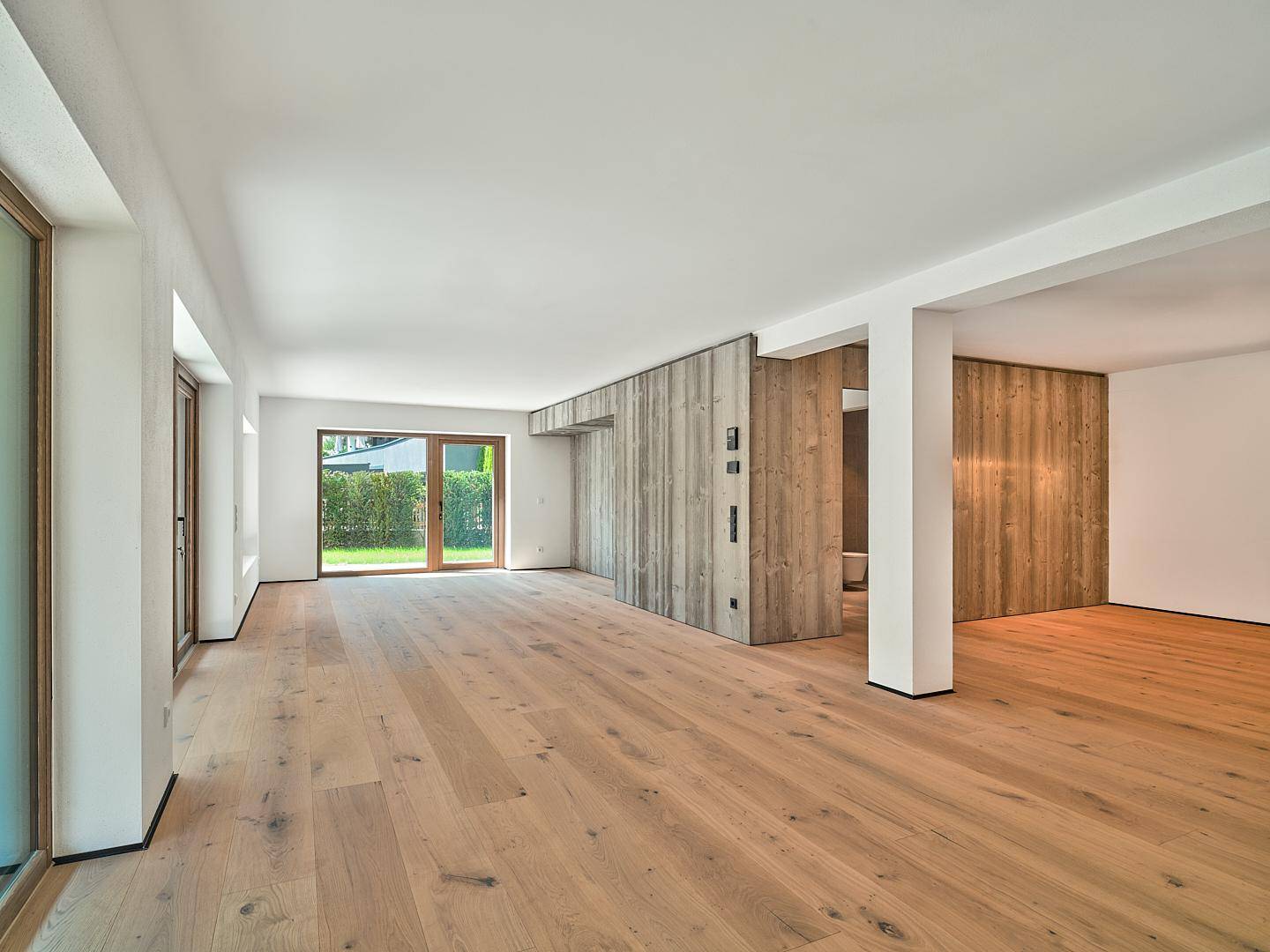 Heller, leerer Wohnraum mit Eichenparkettboden und Zugang zu einem grünen Gartenbereich.
