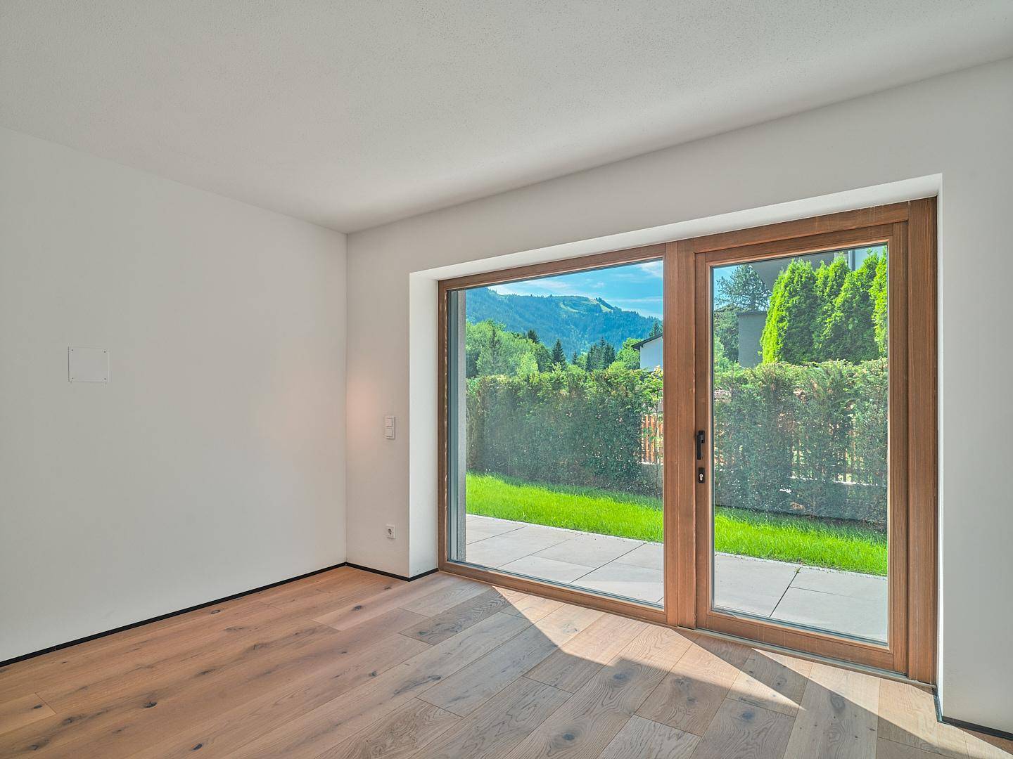Helles Zimmer mit Holzboden und großer Fensterfront, die einen weiten Blick ins Grüne bietet.
