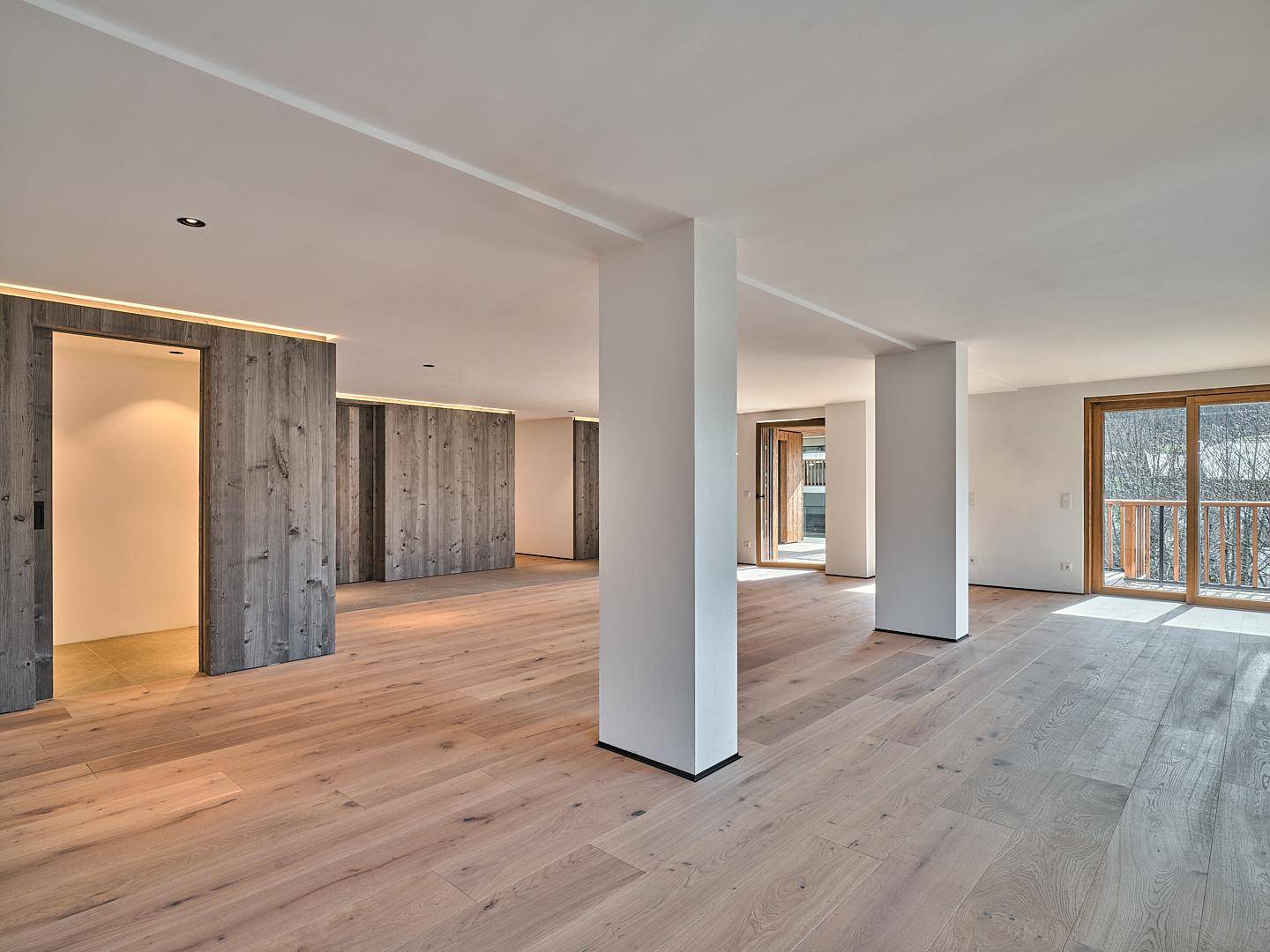 Heller, leerer Wohnbereich mit Holzboden, modernen Säulen und Zugang zu einem Balkon.