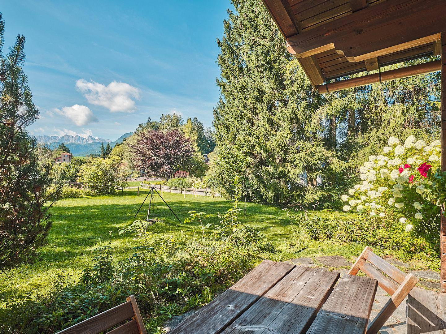Sonniger Außenbereich mit Holztisch und weitem Blick über grüne Wiesen auf die Berge.