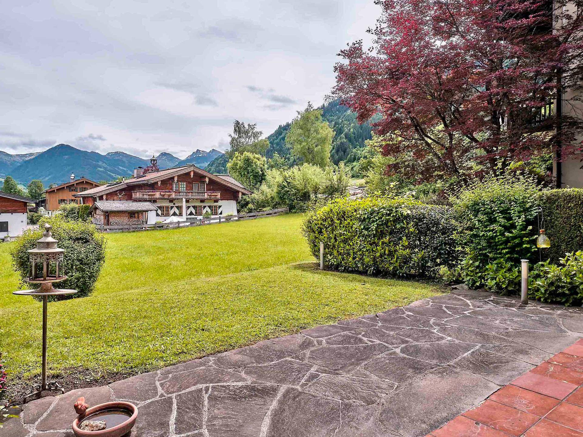 Grüner Gartenbereich mit Rasen, Hecken und traditionellen Häusern vor der beeindruckenden Bergkulisse.