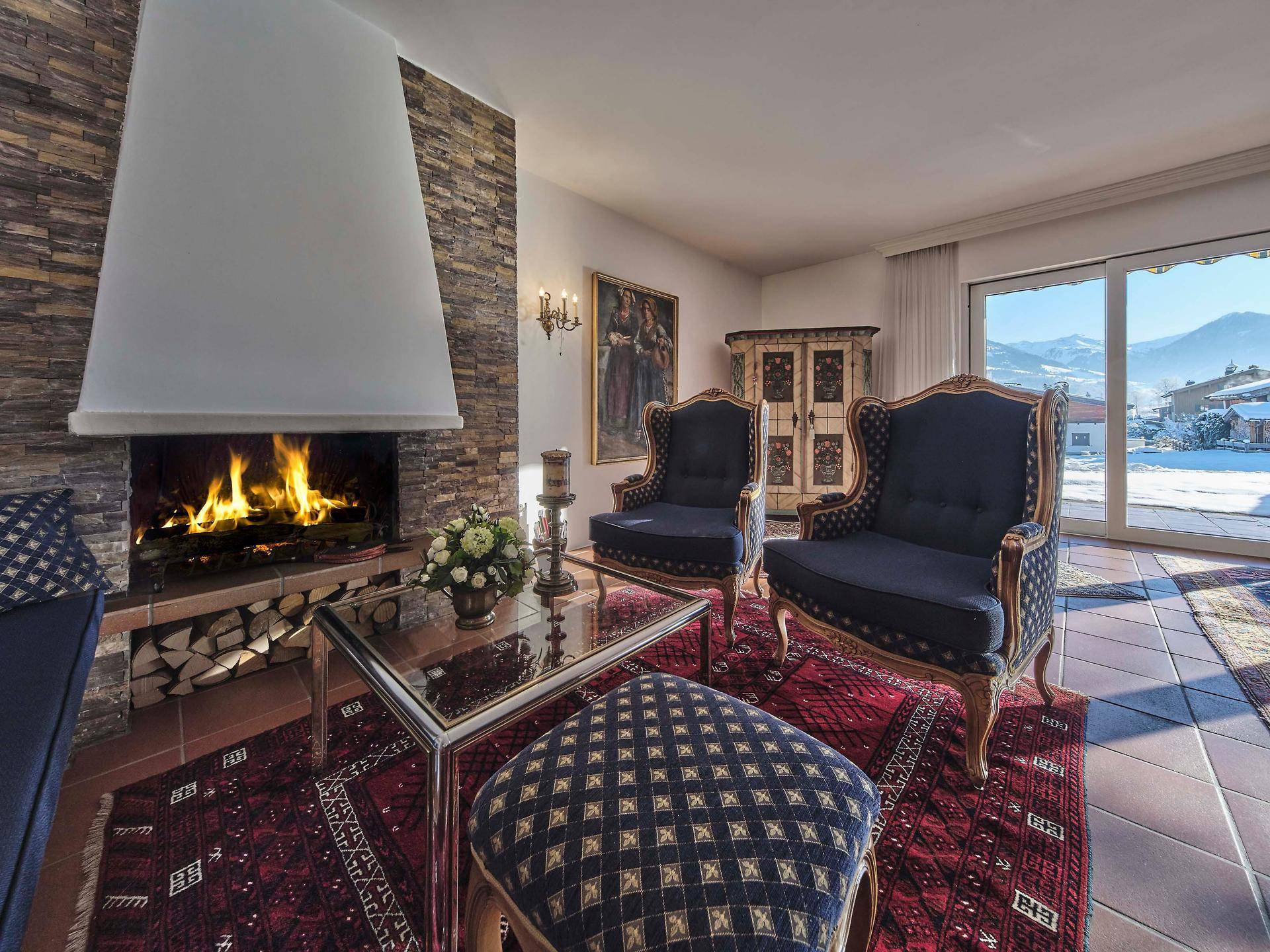 Gemütlicher Wohnbereich mit brennendem Kamin, eleganten blauen Sesseln und Blick auf die verschneiten Berge.