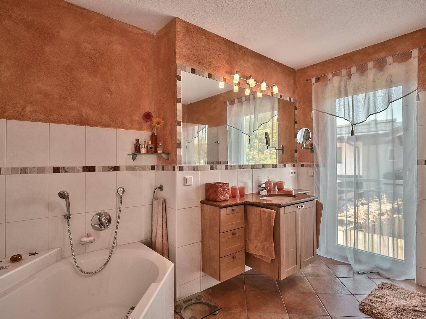 Badezimmer mit Eckbadewanne, großem Spiegel und terracottafarbenen Wänden.