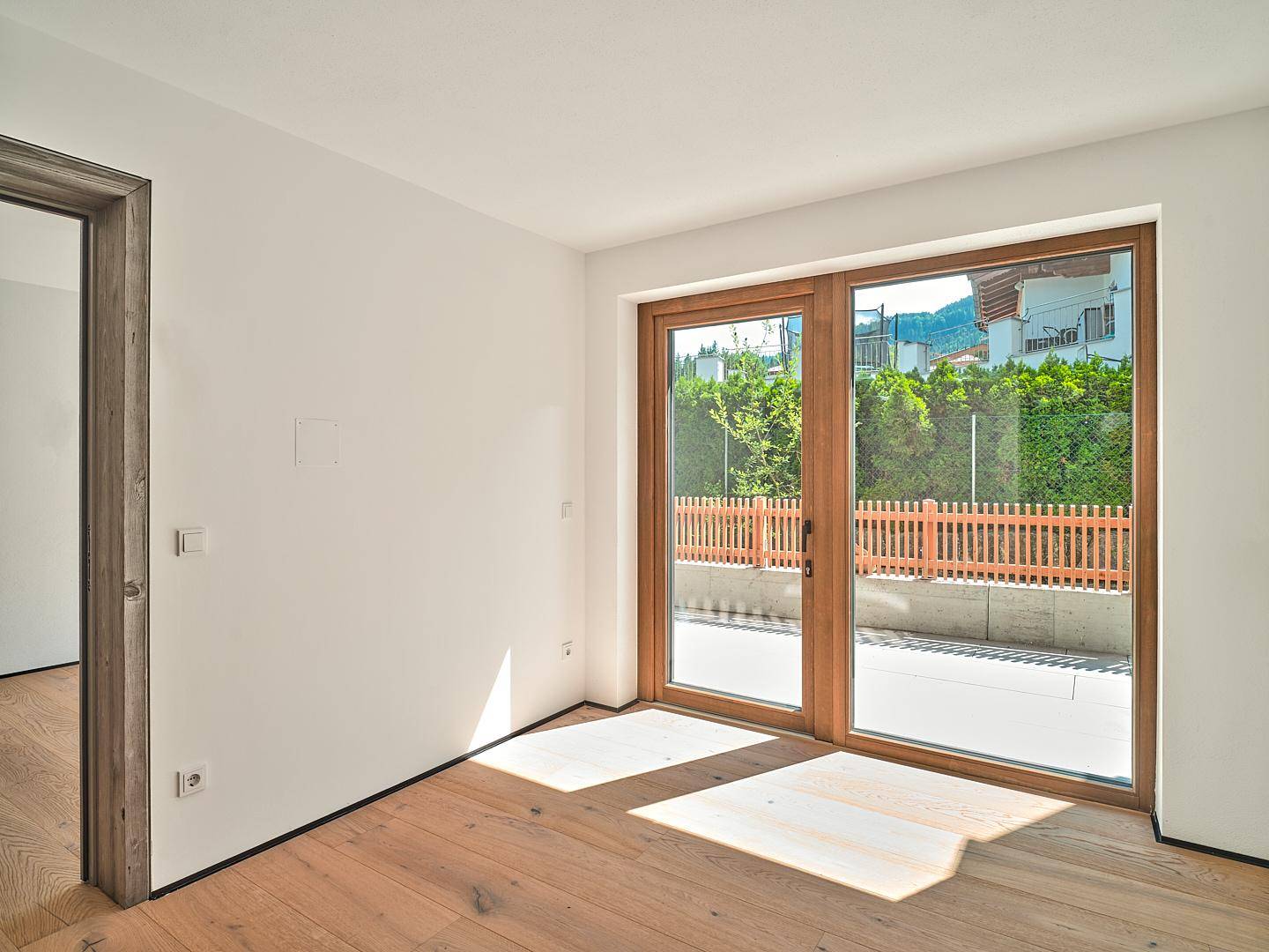 Lichtdurchfluteter, leerer Raum mit Holzboden und großen Fenstern, die Zugang zu einer Terrasse bieten.
