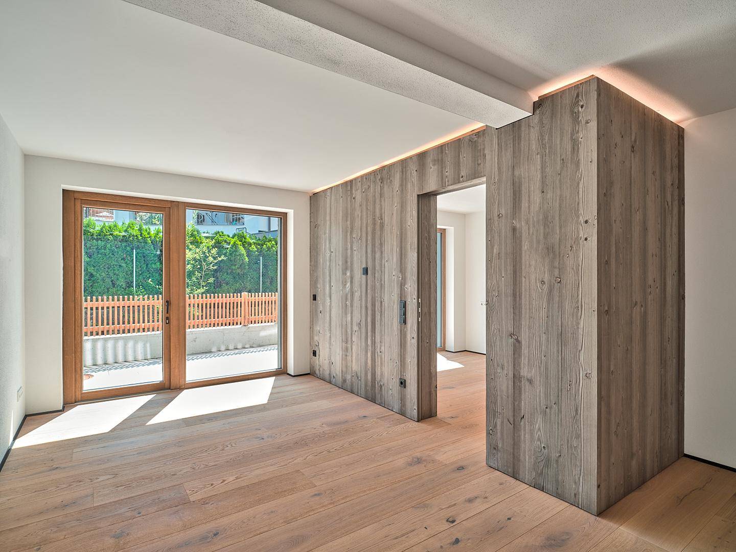 Heller, unmöblierter Raum mit Holzboden, Holzwandelementen und großen Fenstern zum Außenbereich.