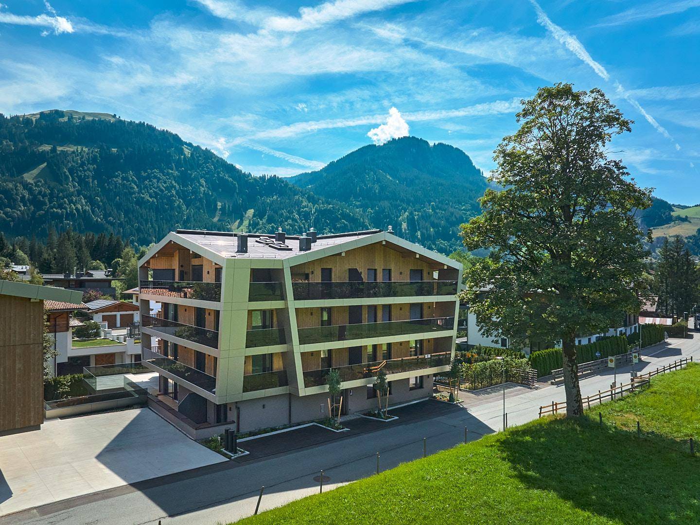Modernes Apartmenthaus in alpiner Landschaft mit Bergen und viel Grün unter strahlend blauem Himmel.