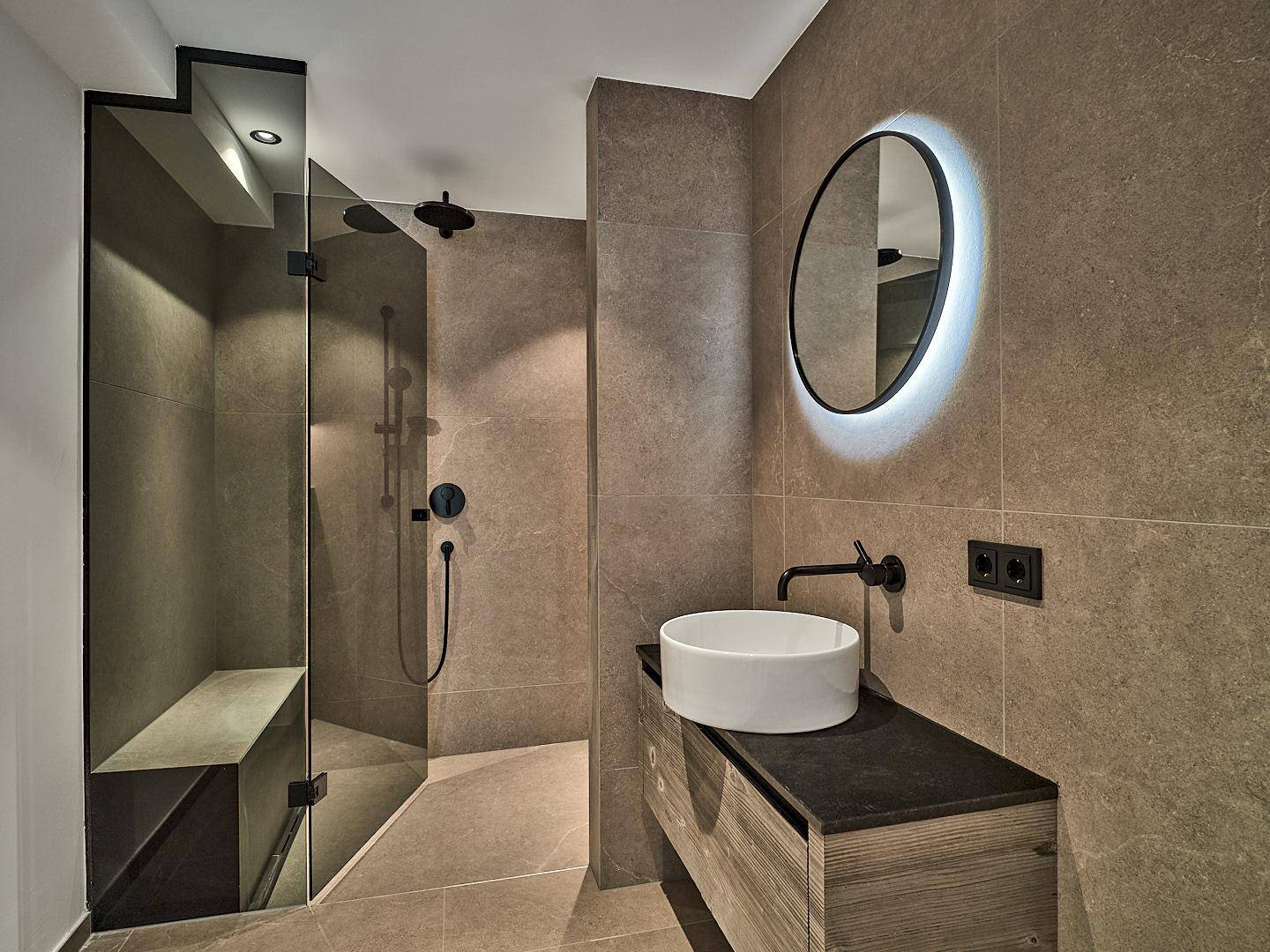 Modernes Badezimmer mit begehbarer Dusche, runden Spiegel und dunklen Fliesen in zeitgemäßem Design.