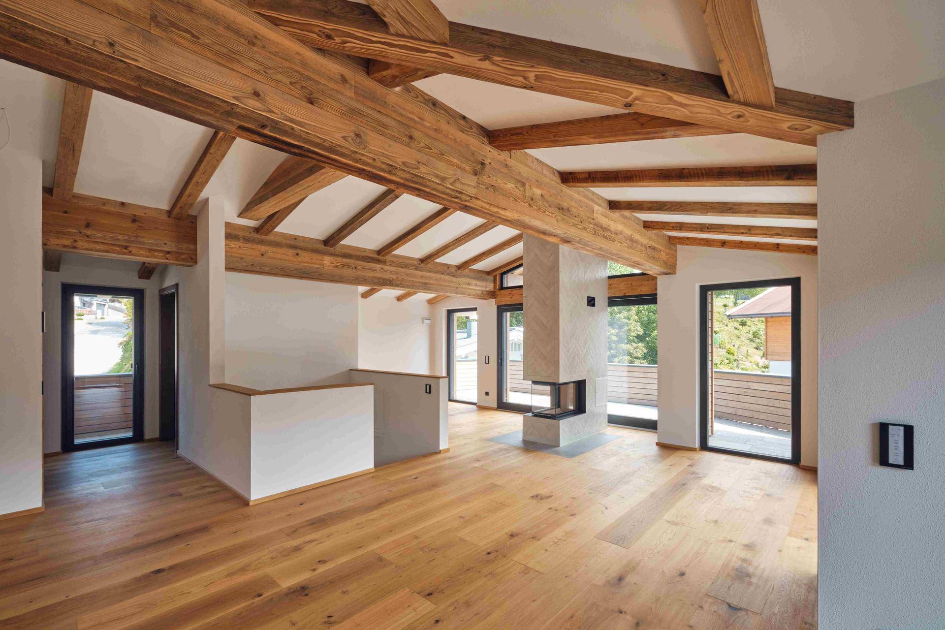 Geräumiger Wohnbereich mit sichtbaren Holzbalken, elegantem Kamin und viel Tageslicht durch große Fenster.