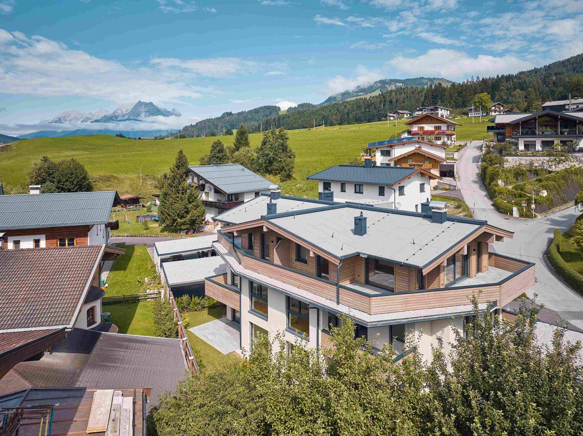 Luftaufnahme eines modernen Wohnkomplexes in alpiner Landschaft mit grünen Hügeln und Bergen.