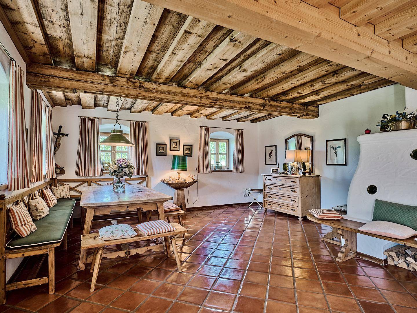 Rustikaler Wohn-Essbereich mit Holzbalkendecke, Terrakottafliesen und traditionellem Kachelofen.