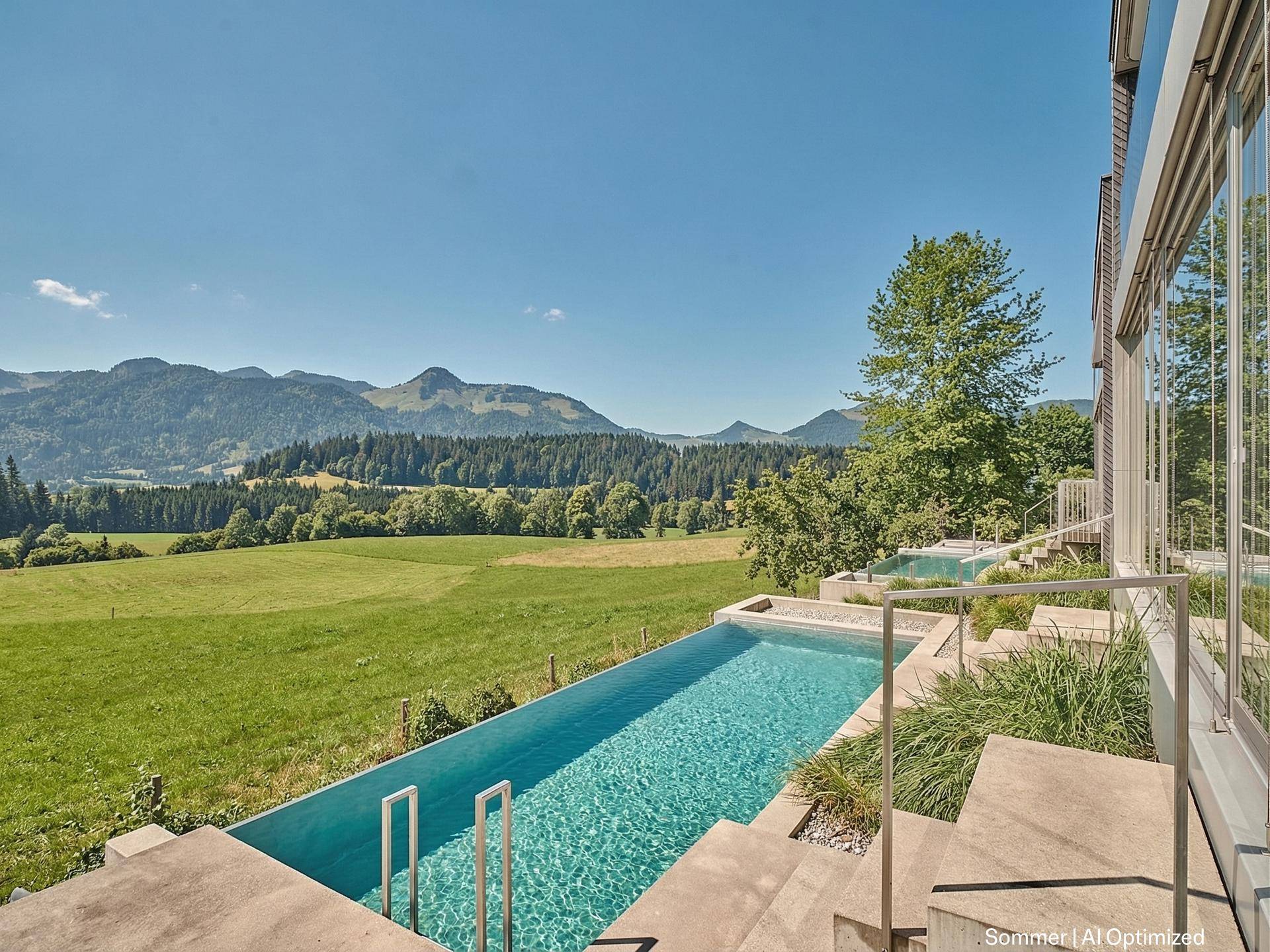 Außenpool mit Bergblick, umgeben von grünen Wiesen und Bäumen unter strahlend blauem Himmel.